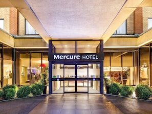 The entrance to the Mercure Hotel with a glass facade, a sign above the doors, and a tiled pavement leading up to the entrance.