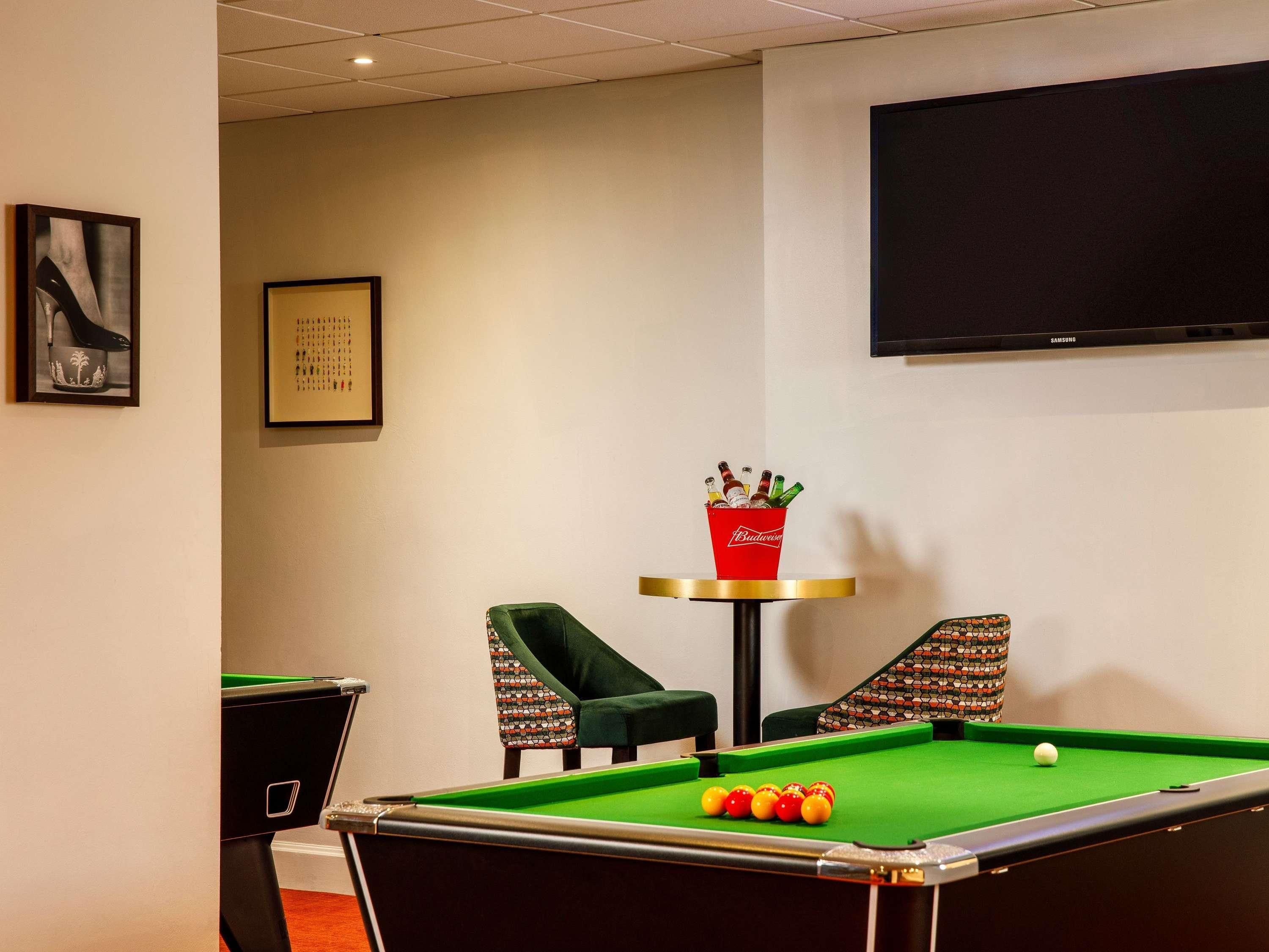 A pool table with balls, a red bucket with bottles, two patterned chairs, a framed picture, and a television on the wall.