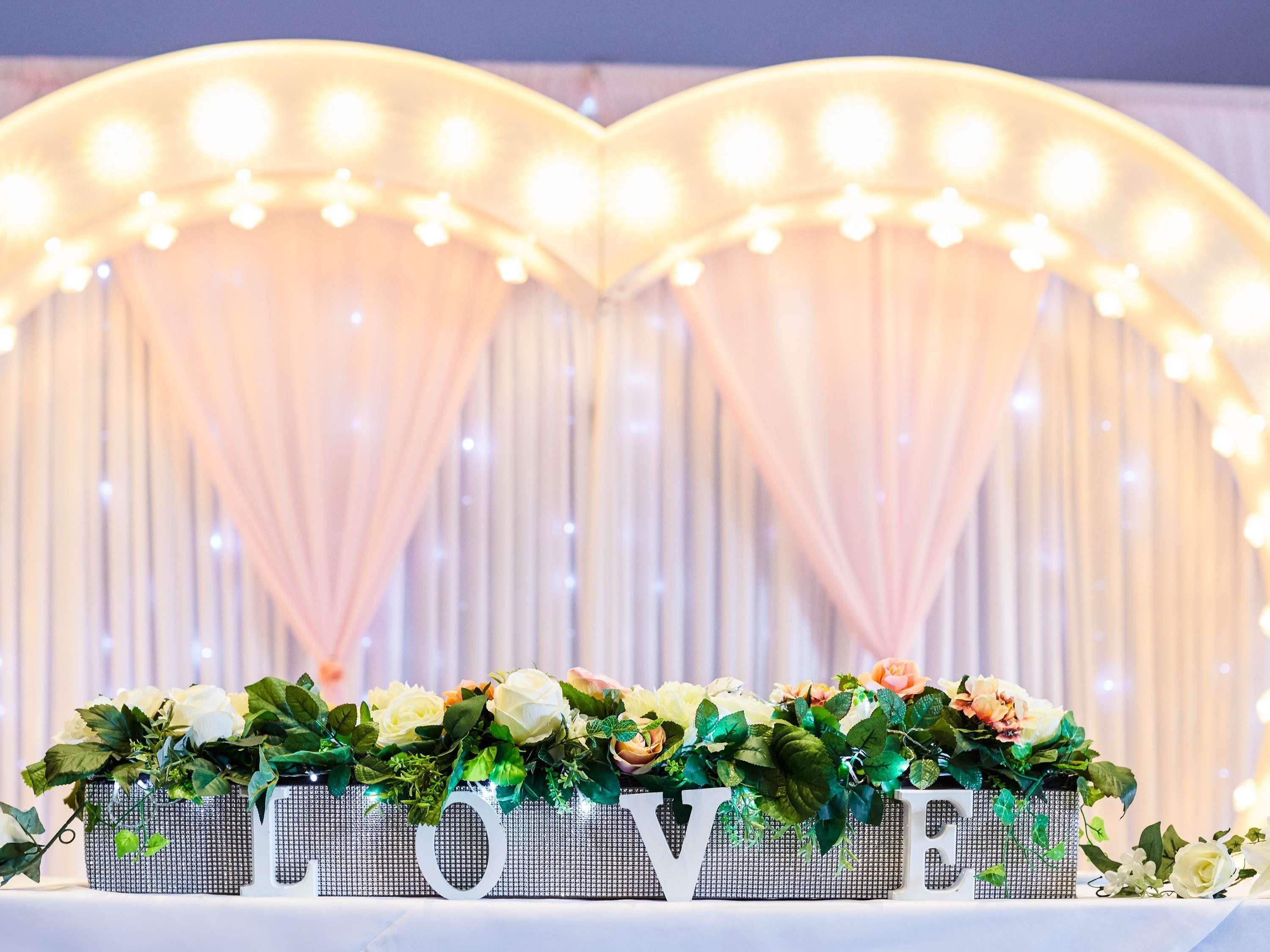 Un escenario con un fondo de tela drapeada y luces de cuerda, decorado con arreglos florales y un letrero iluminado de gran tamaño.