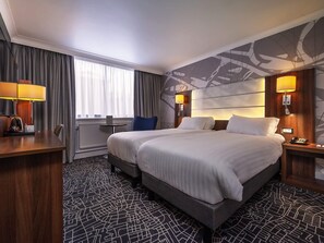 A hotel room with a large bed, two bedside tables with lamps, a desk with a chair, a small table, and a window with curtains.