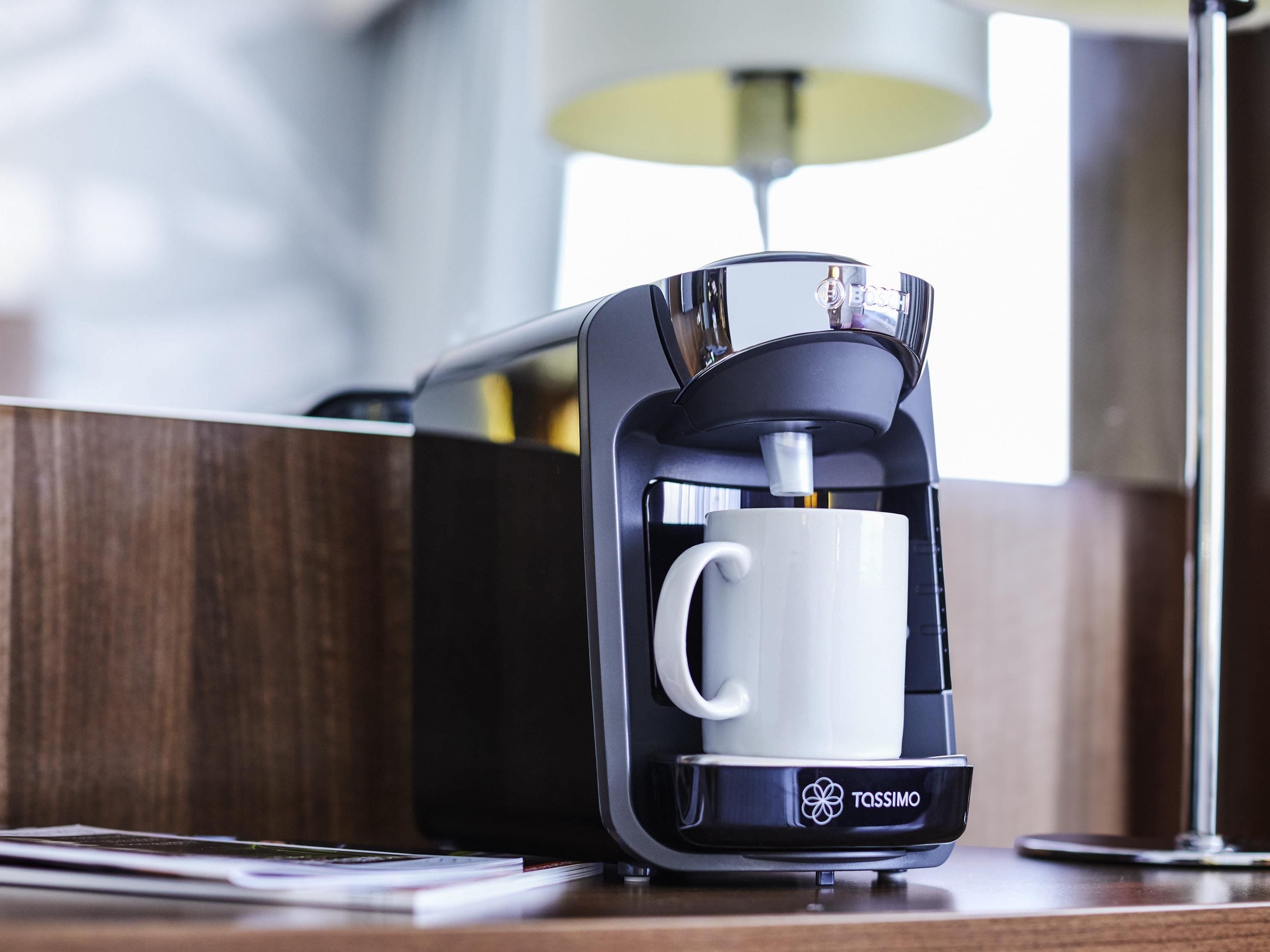 A Tassimo coffee machine with a white mug on its base, placed on a wooden surface with a lamp in the background.