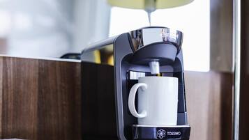 A Tassimo coffee machine with a white mug on its base, placed on a wooden surface with a lamp in the background.