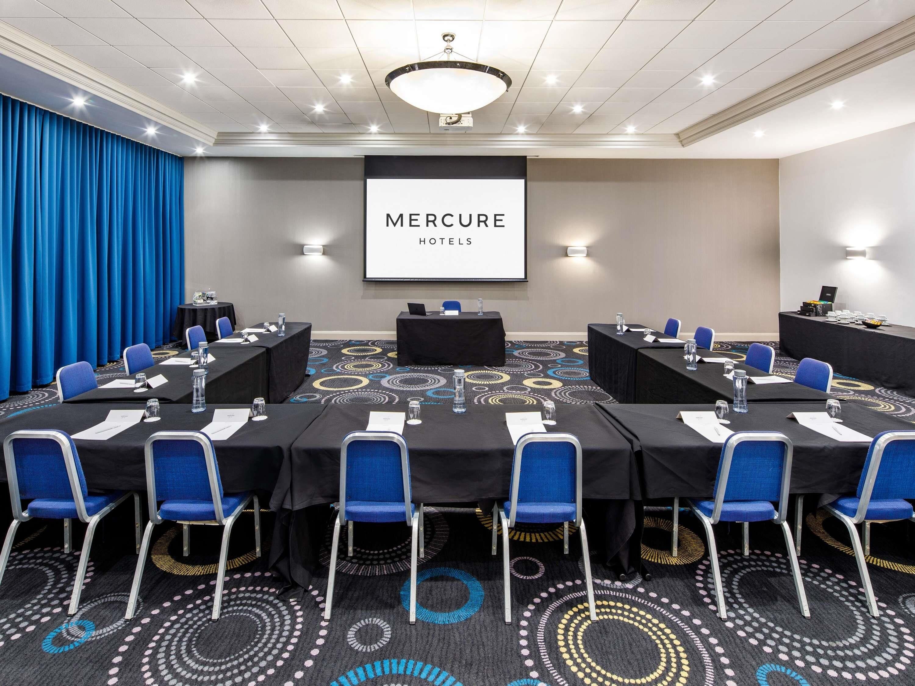 A conference room with a large screen displaying 'MERCURE HOTELS,' tables with chairs, and a patterned carpet.