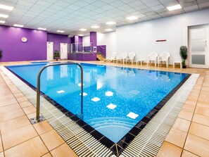 A swimming pool with a tiled deck, a clock on the wall, and white chairs.