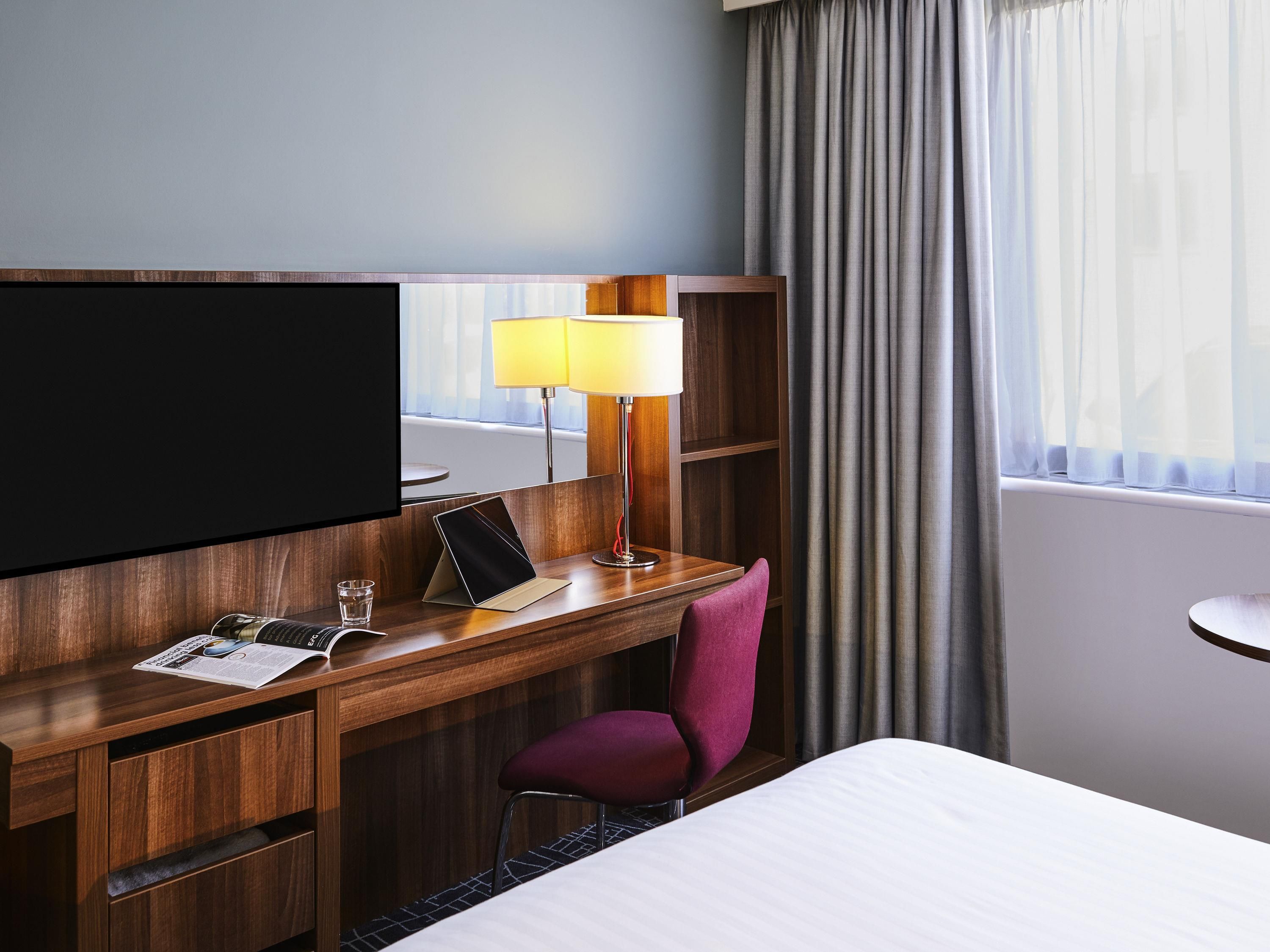 A hotel room with a bed, a desk with a laptop, a TV, a lamp, and a chair.