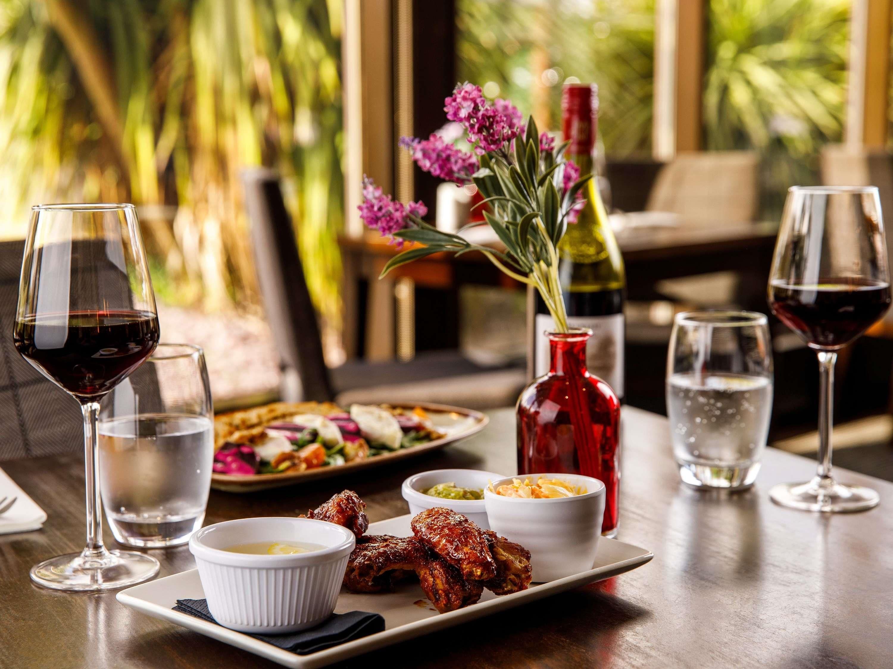 A dining table with a plate of grilled chicken wings, a red vase with flowers, and several glasses of wine.