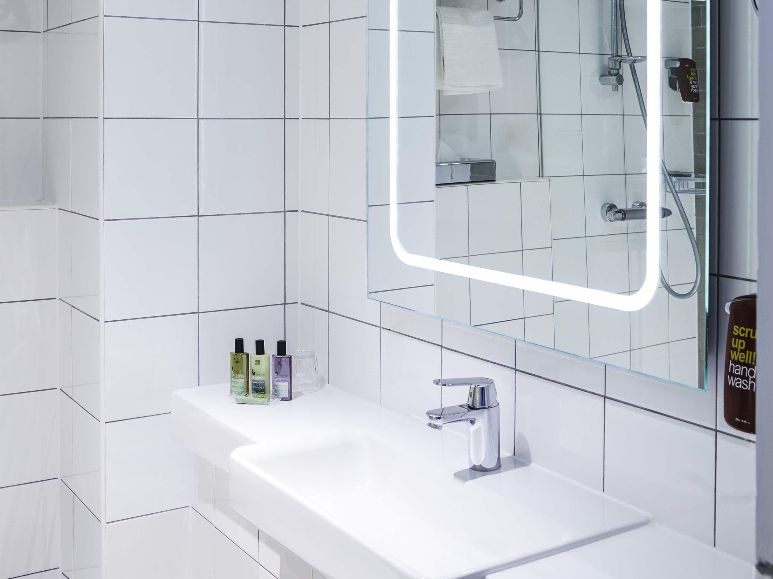 Ein modernes Badezimmer mit weißen Fliesen, einer Glasablage für Toilettenartikel, einem Spiegel mit Beleuchtung und einer Duschkabine.