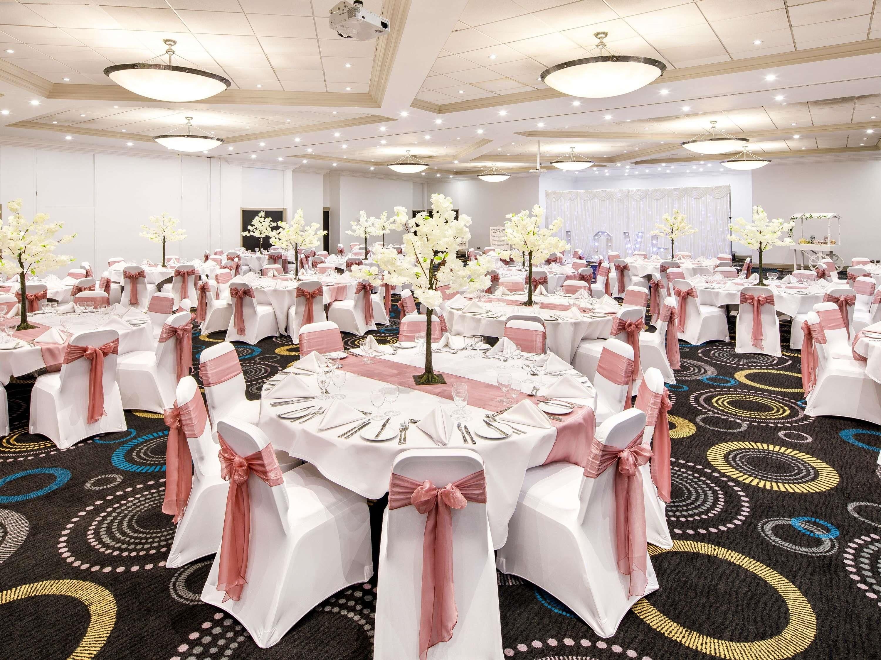 A banquet hall with round tables, white chairs with pink sashes, and floral centerpieces.