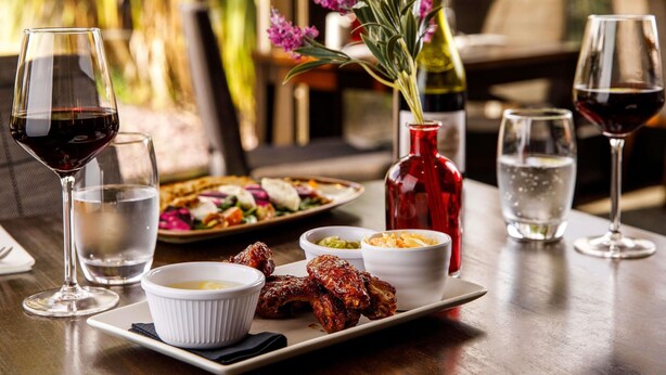 Ein Esstisch mit einem Teller gegrillten Hähnchenflügeln, einer roten Vase mit Blumen und mehreren Weingläsern.
