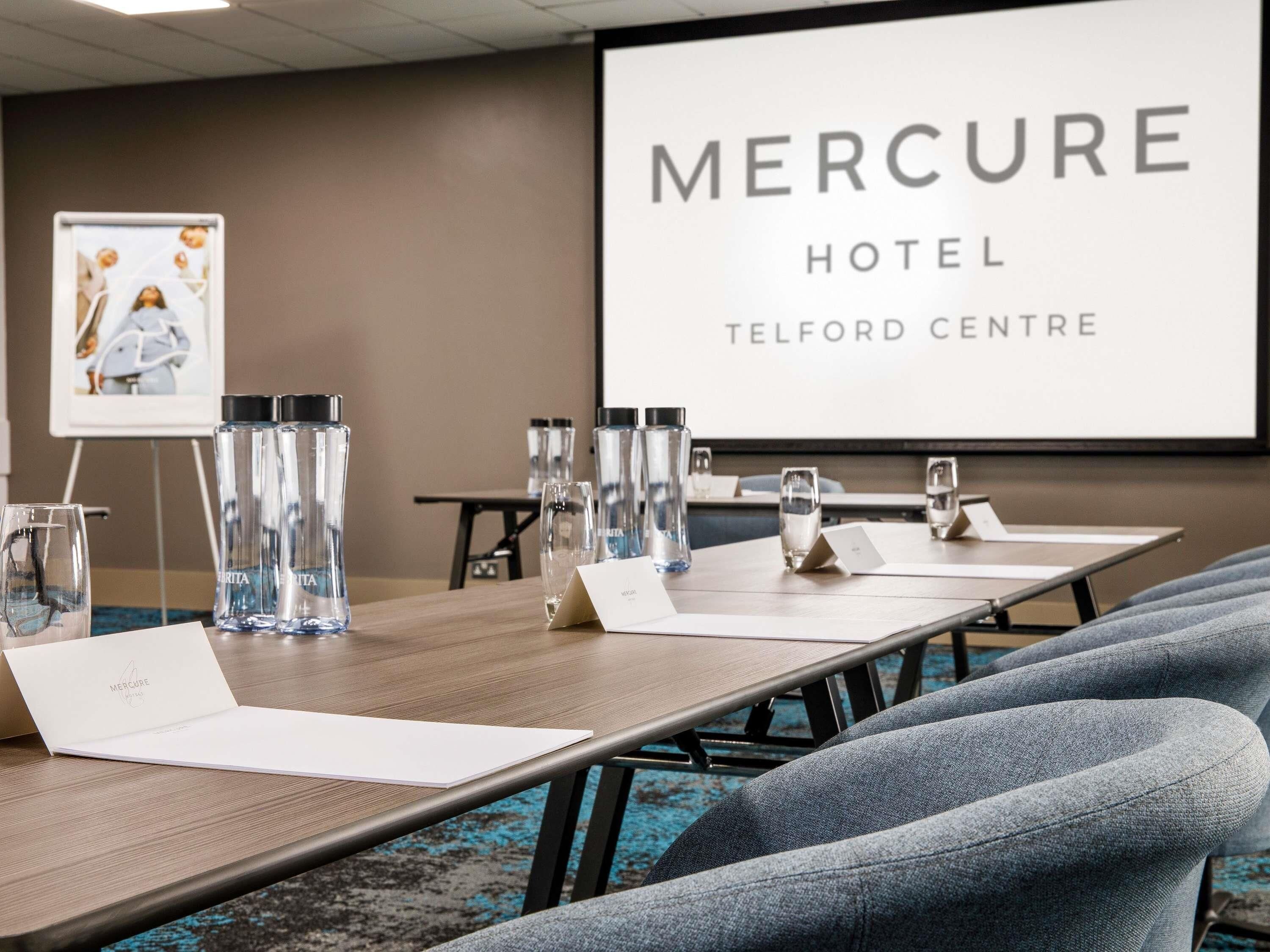 A conference room with a large screen displaying 'Mercure Hotel Telford Centre,' a wooden table with water bottles, glasses, and name cards, and grey upholstered chairs.