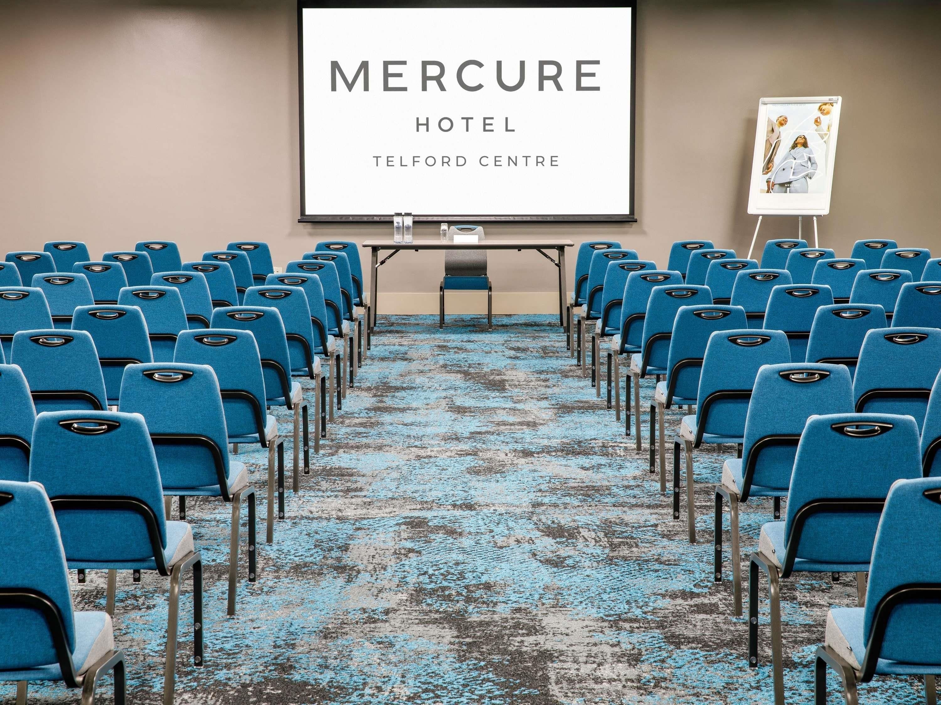 Una sala conferenze con file di sedie blu, uno schermo di proiezione e un cartello con la scritta 'Mercure Hotel, Telford Centre'.