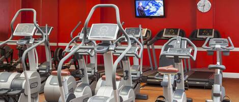 A gym with various cardio machines, a television on the wall, and a clock.