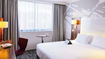 A hotel room with a large bed, a desk with a lamp, a chair, a small table, and a window with curtains.
