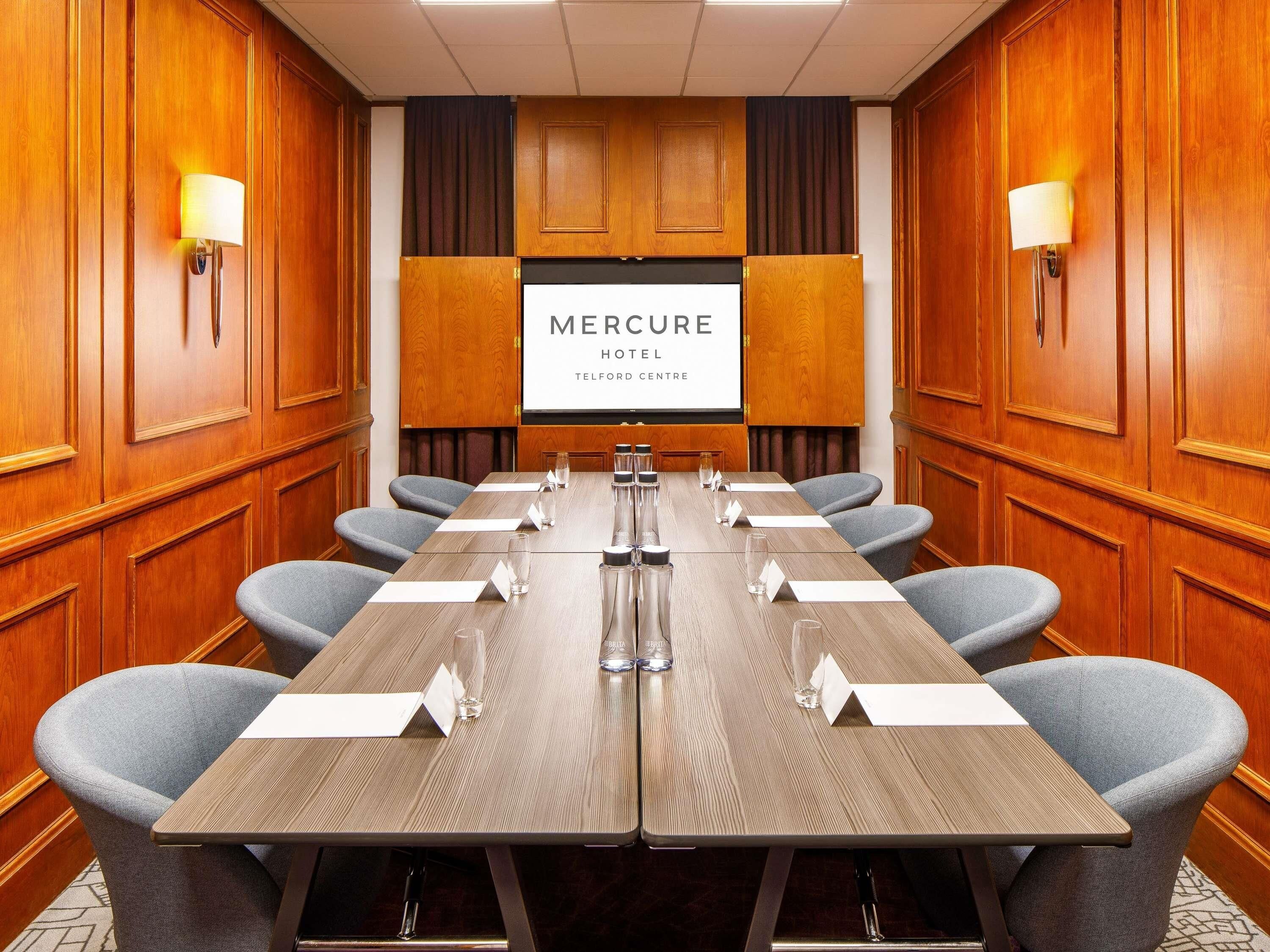 Un salón de conferencias con una larga mesa, sillas, tarjetas de identificación y una pantalla mostrando 'Mercure Hotel Telford Centre'.