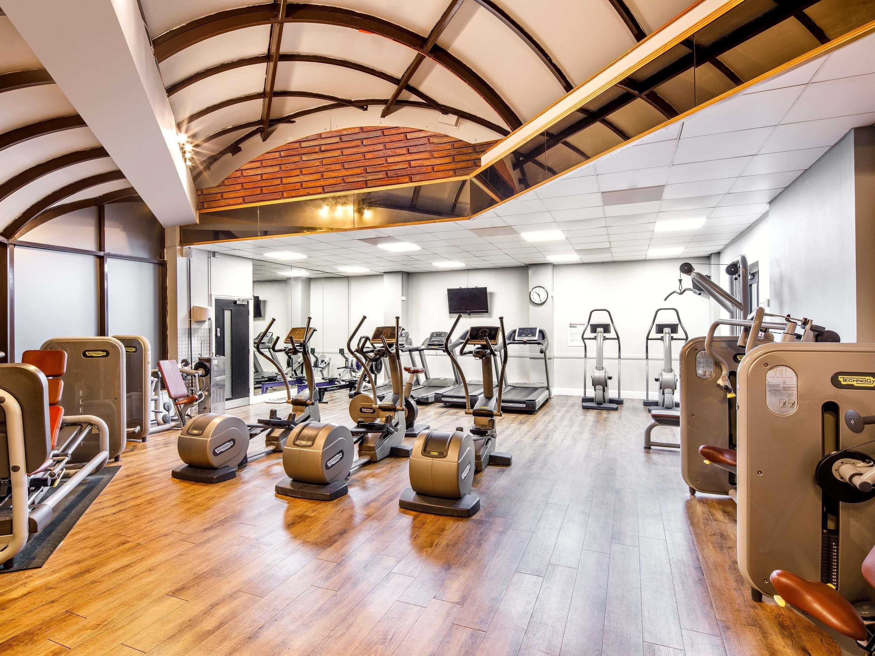 A gym with various exercise machines, wooden flooring, and a brick wall.