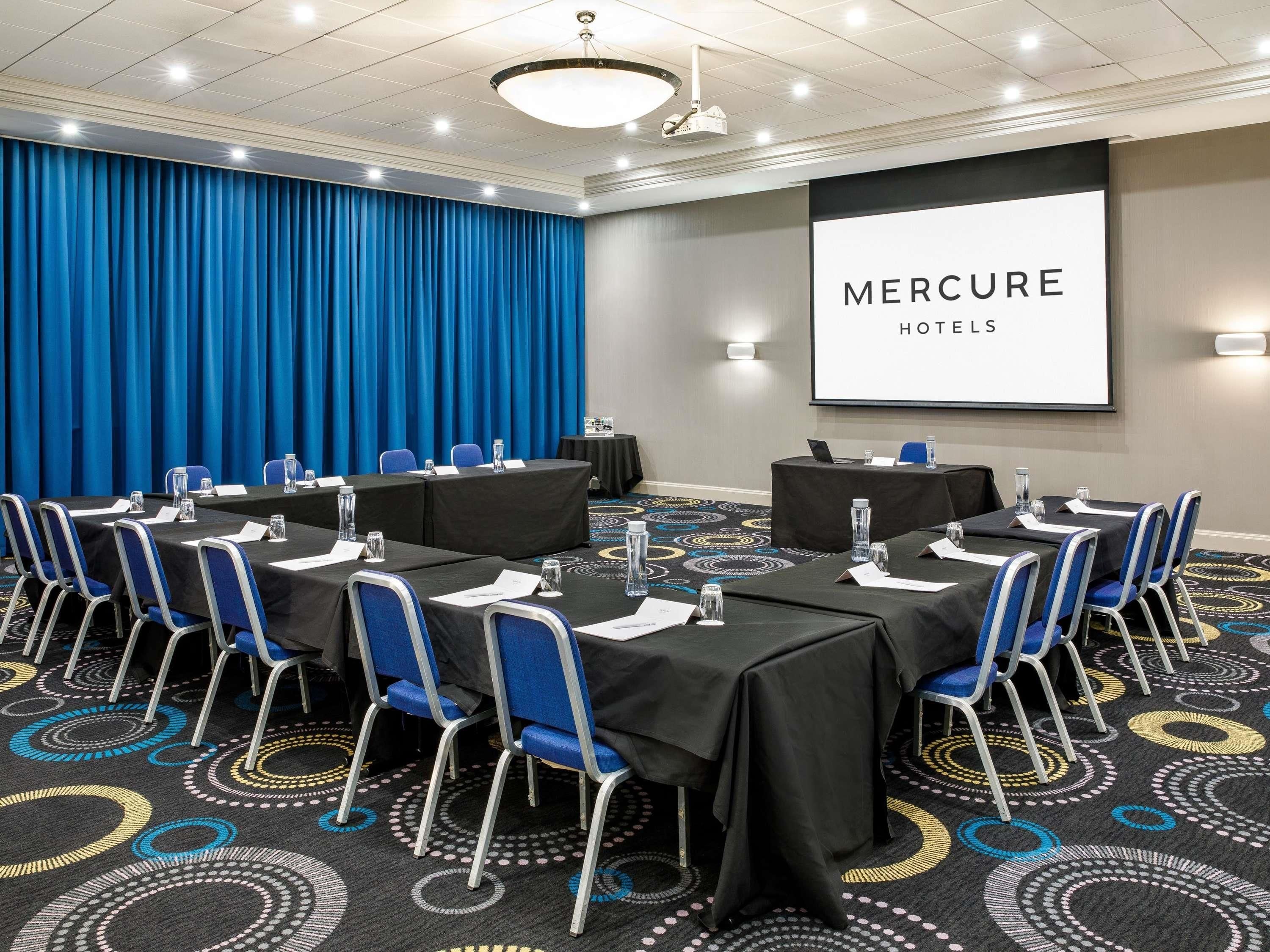 A conference room with a large screen displaying 'MERCURE HOTELS' and a blue curtain backdrop.