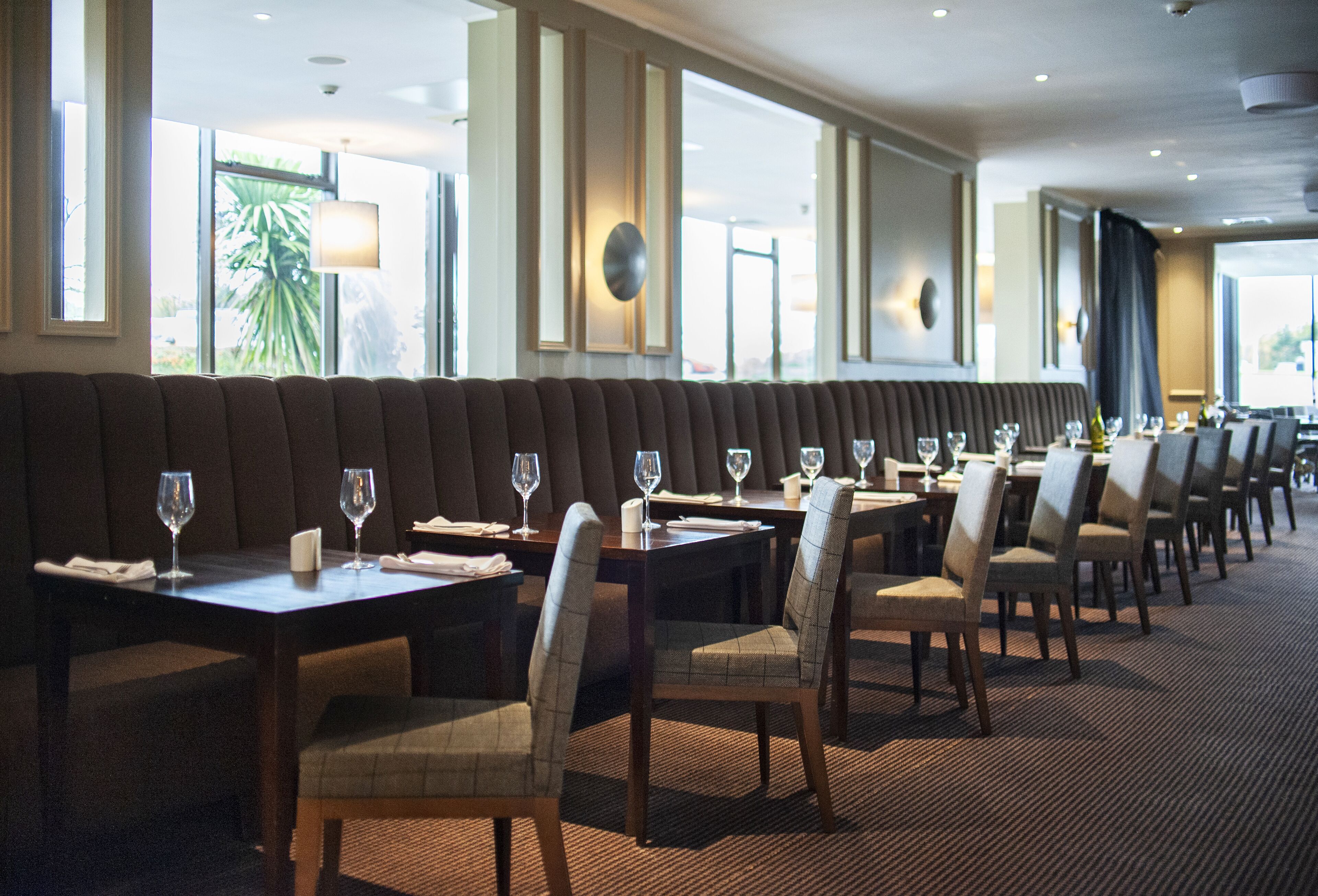 A restaurant with a long bench seating area, tables with chairs, and large windows with views of palm trees.