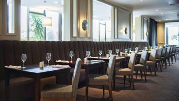 A restaurant with a long bench seating area, tables with chairs, and large windows with views of palm trees.