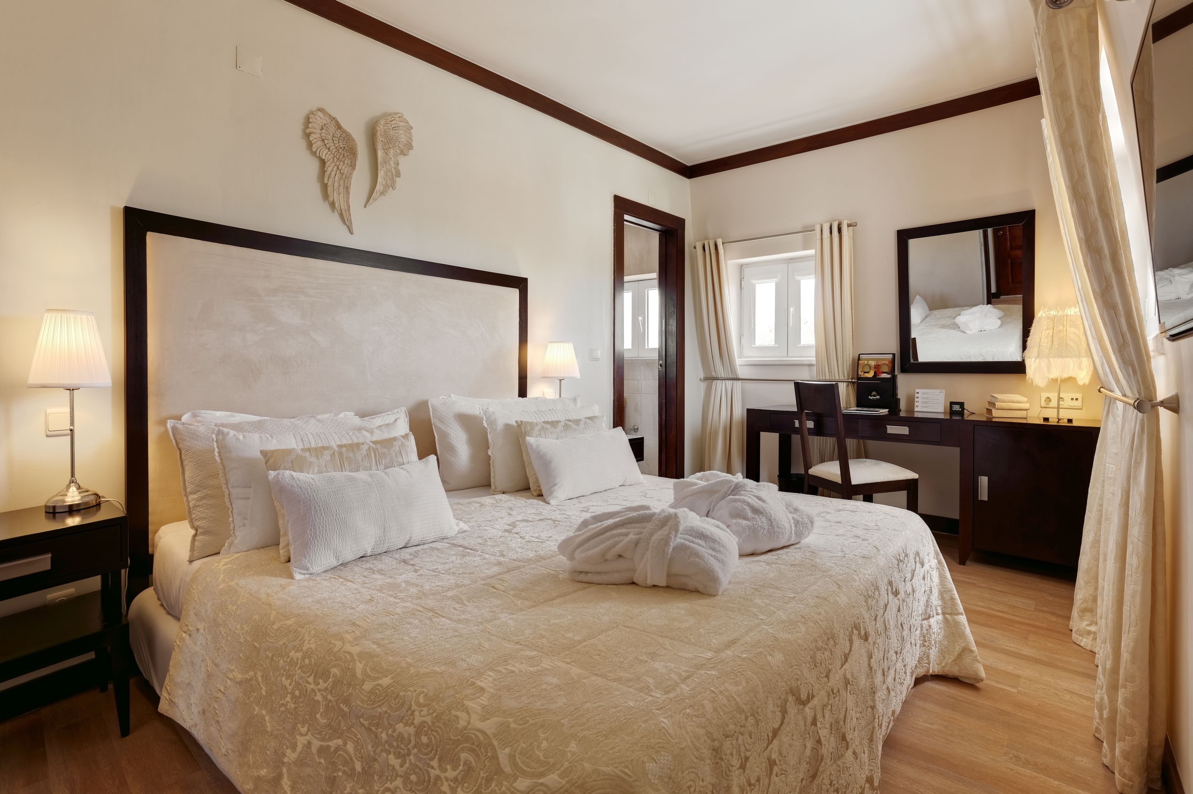 A hotel room with a large bed, two bedside lamps, a desk with a chair, a mirror, and a wardrobe.
