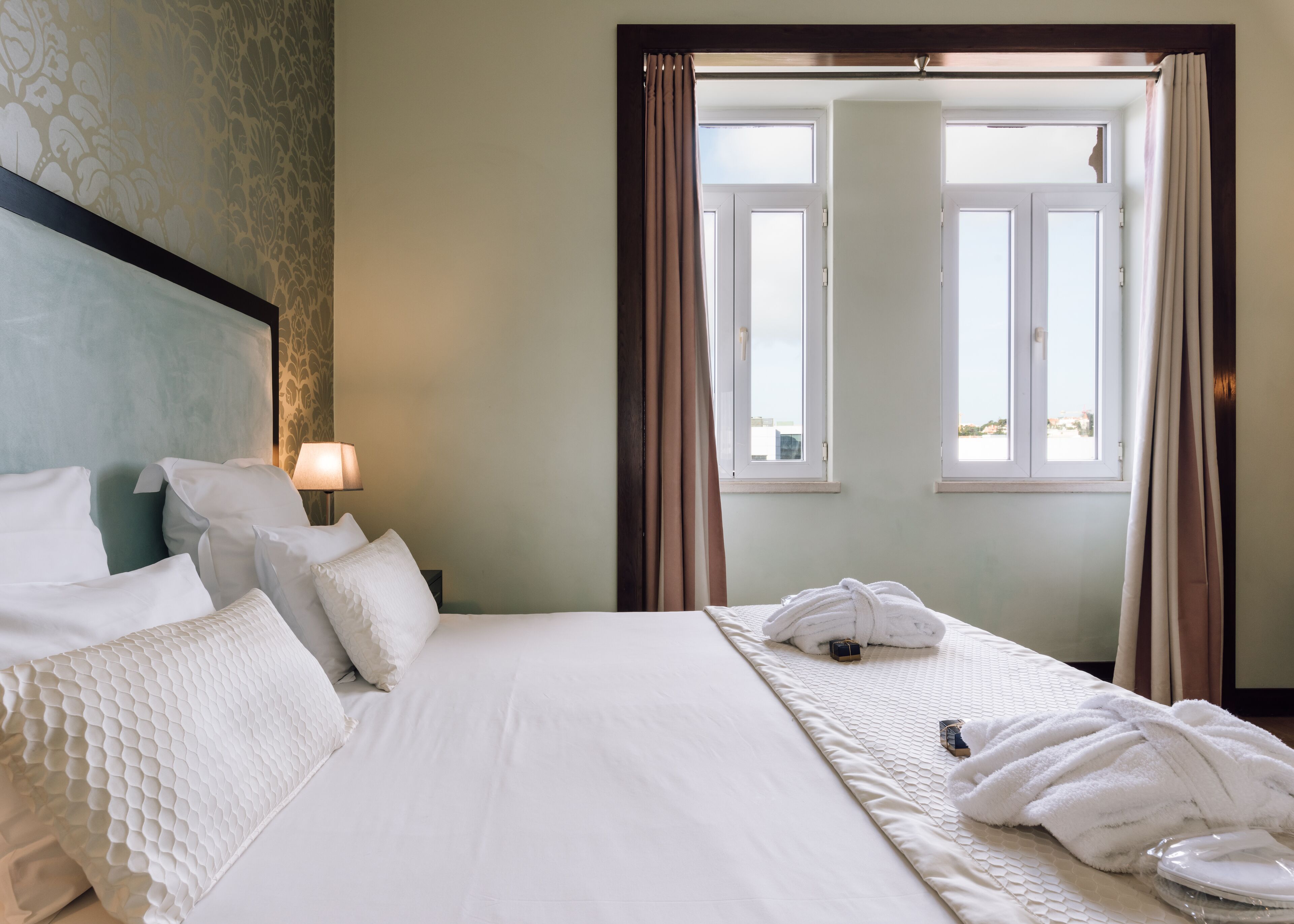 Una habitación de hotel con una cama grande, ropa de cama blanca, cabecero, mesita de noche con lámpara y vistas a través de una ventana con cortinas.