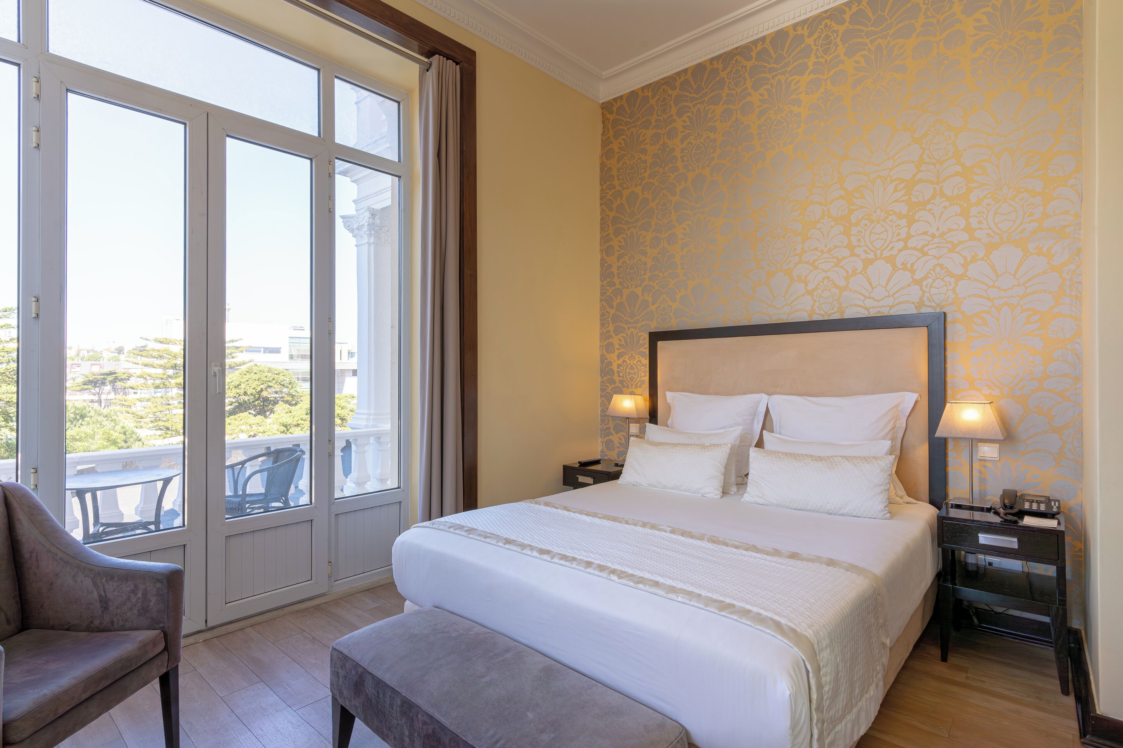 Habitación de hotel con una cama grande, una silla, un banco, una mesita de noche con lámpara y vistas a través de un ventanal.