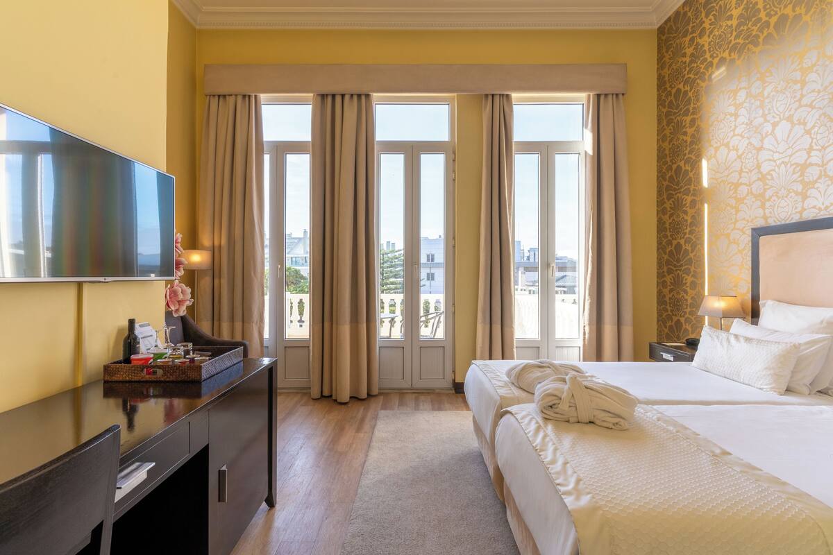 A hotel room with a large bed, a desk with a flat-screen TV, a dresser, and a view of the city through the windows.