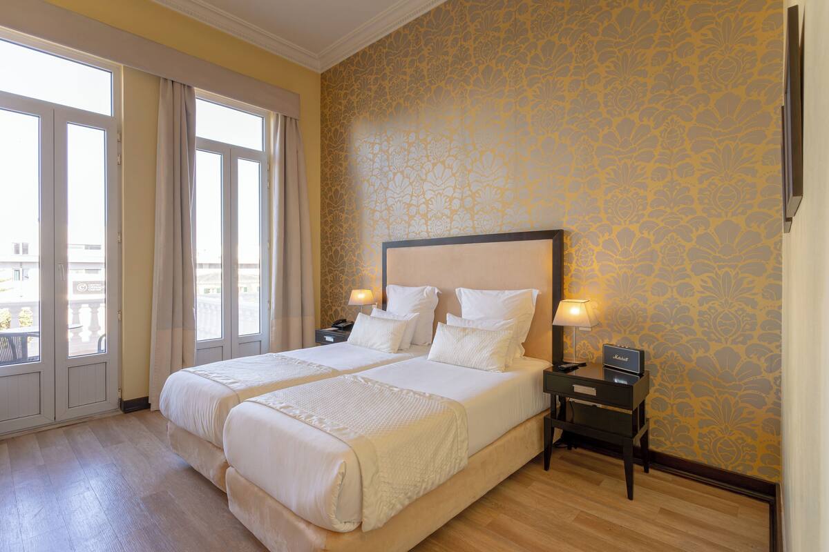 A hotel room with two beds, a nightstand with a lamp, and a view through the window.
