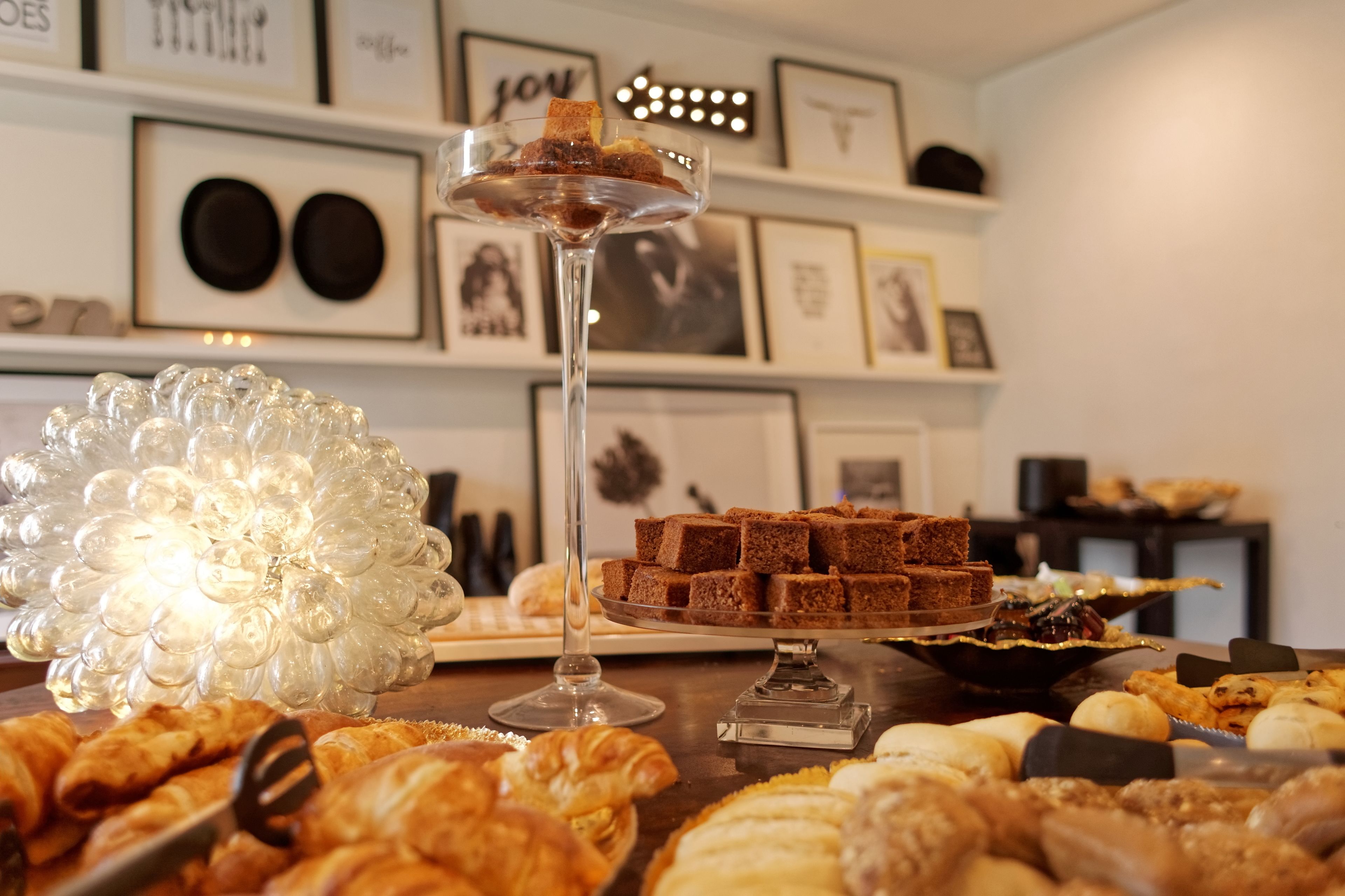 A buffet table with various pastries and desserts, including croissants and brownies, in a well-lit room with framed pictures on the wall.