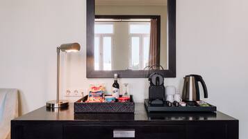 A hotel desk with a lamp, a coffee maker, a tray with snacks, and a mirror above.