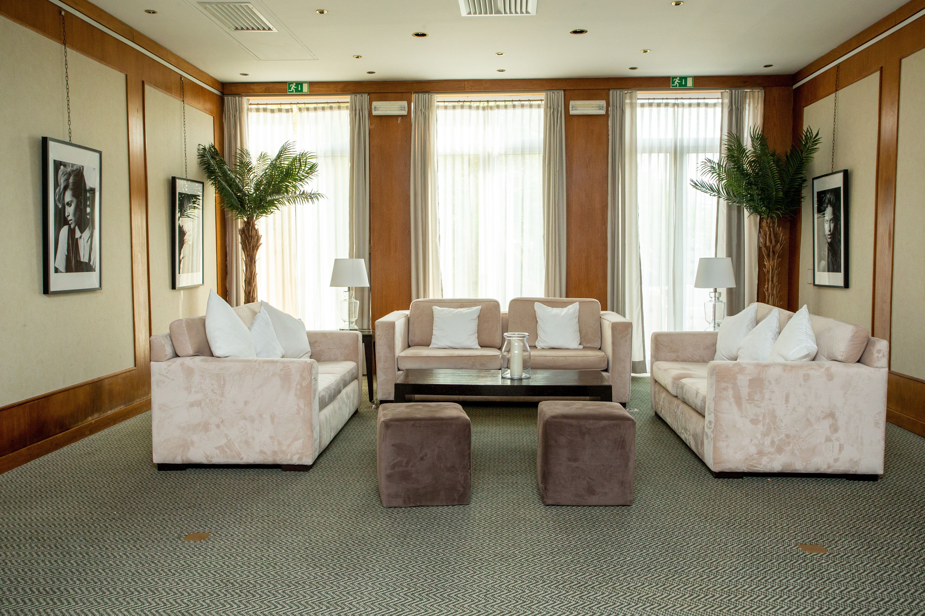 A hotel lobby with two sofas, a coffee table, and two ottomans. There are two potted plants and framed pictures on the walls.