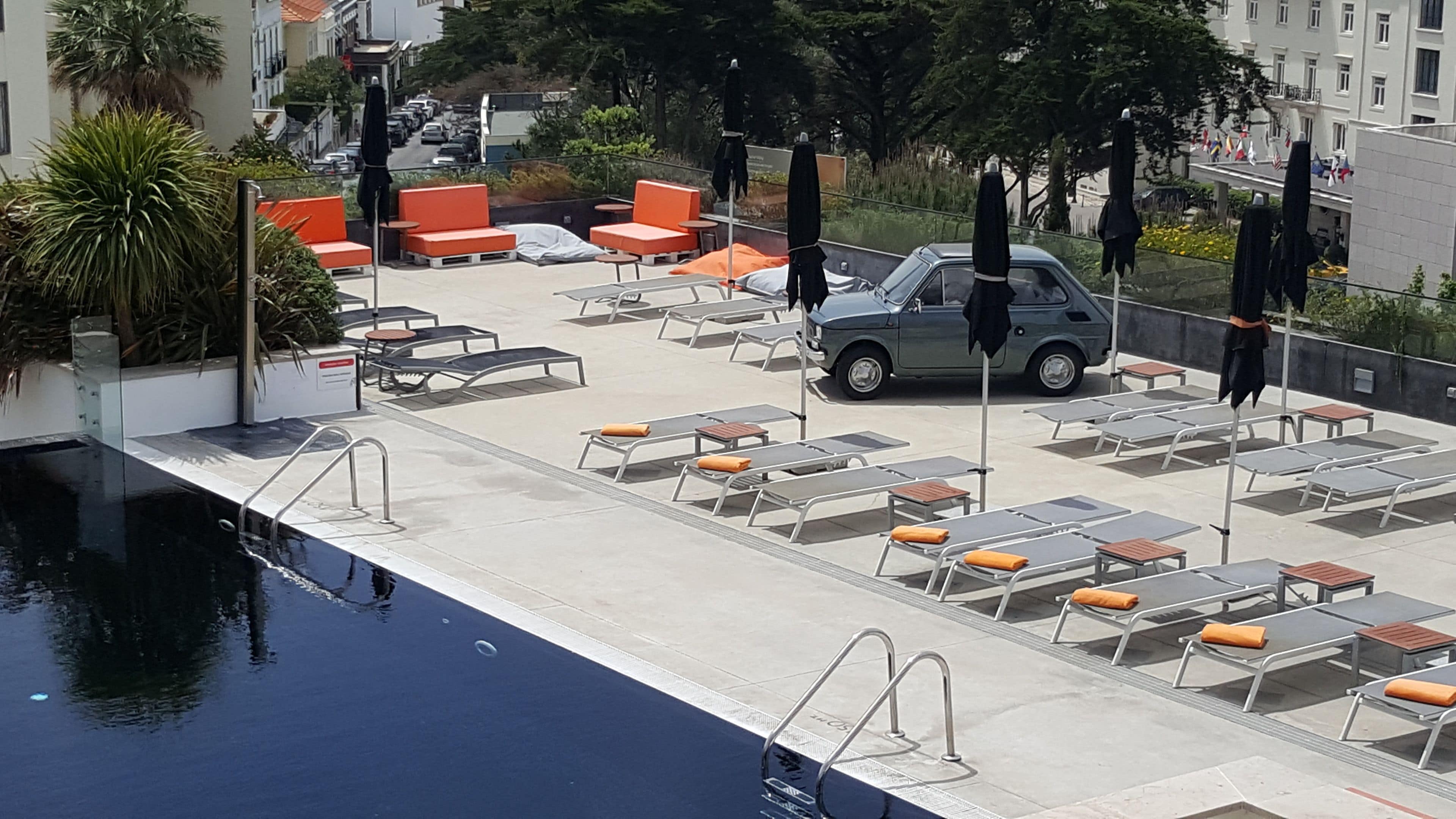 Un área de piscina en la azotea con tumbonas, un coche y un edificio al fondo.