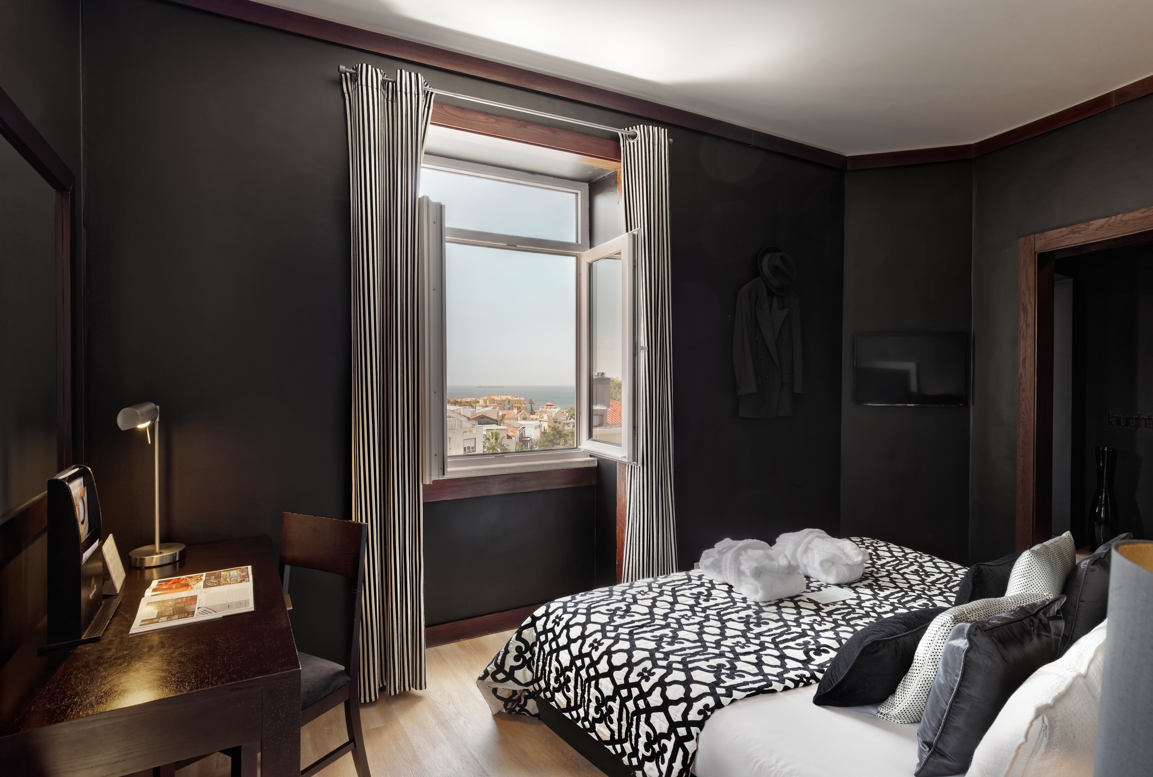 A modern hotel room with a large window, a desk with a lamp, a bed with patterned bedding, and a view of the city.