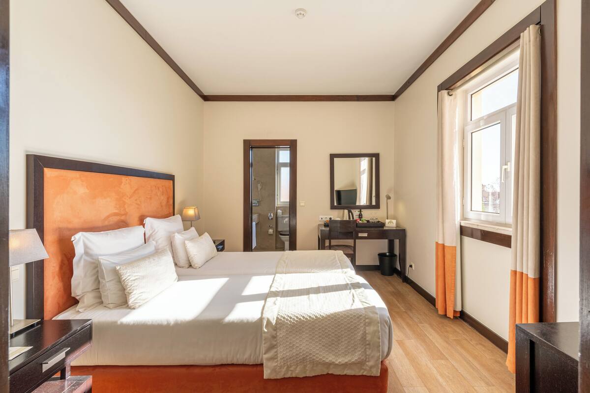 A hotel room with a large bed, a desk, a mirror, and a window with curtains.