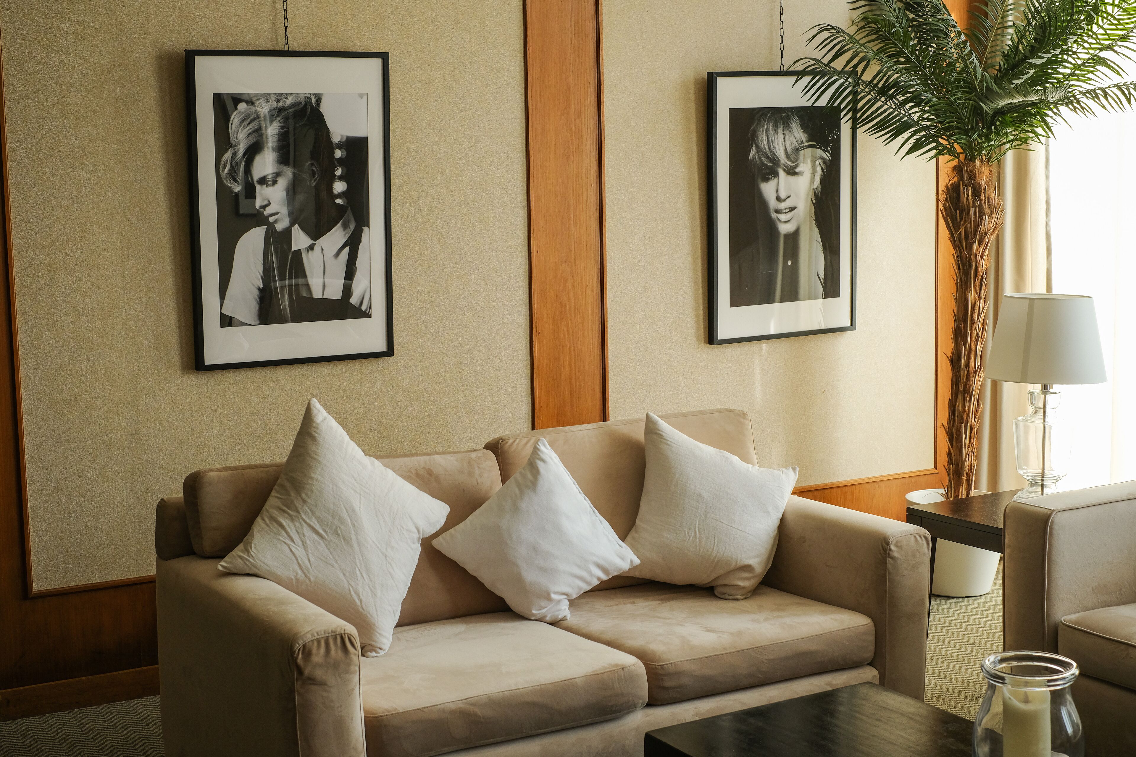 A beige sofa with white cushions, a small table, a lamp, and two framed pictures on the wall.