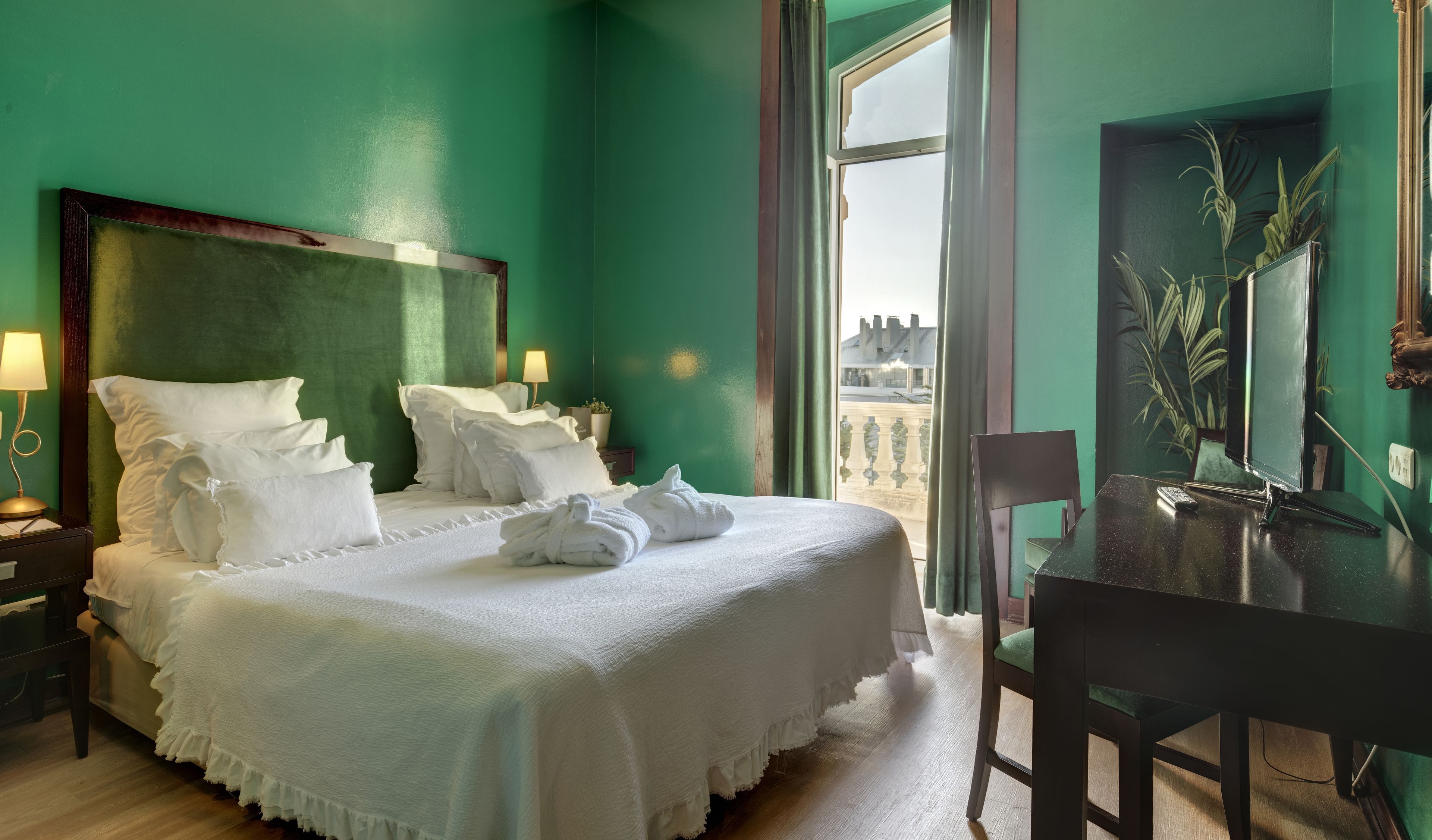Una cama bien hecha con sábanas y almohadas blancas, una mesita de noche con lámpara, y vista a la ciudad a través de un ventanal grande.