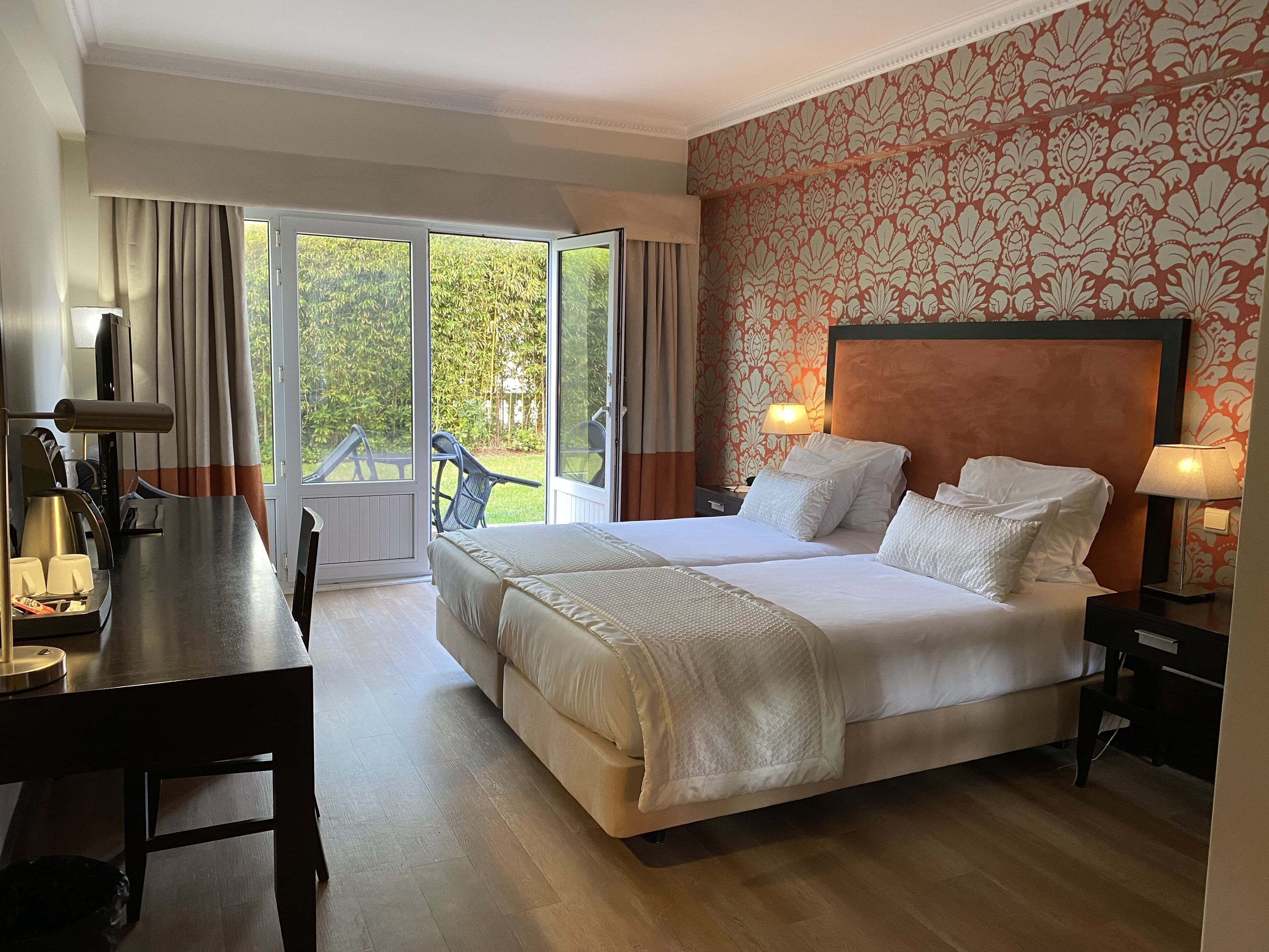 Habitación de hotel con una cama grande, un escritorio con una silla, una mesa con tazas y una tetera, y vistas al exterior a través de una puerta corredera.