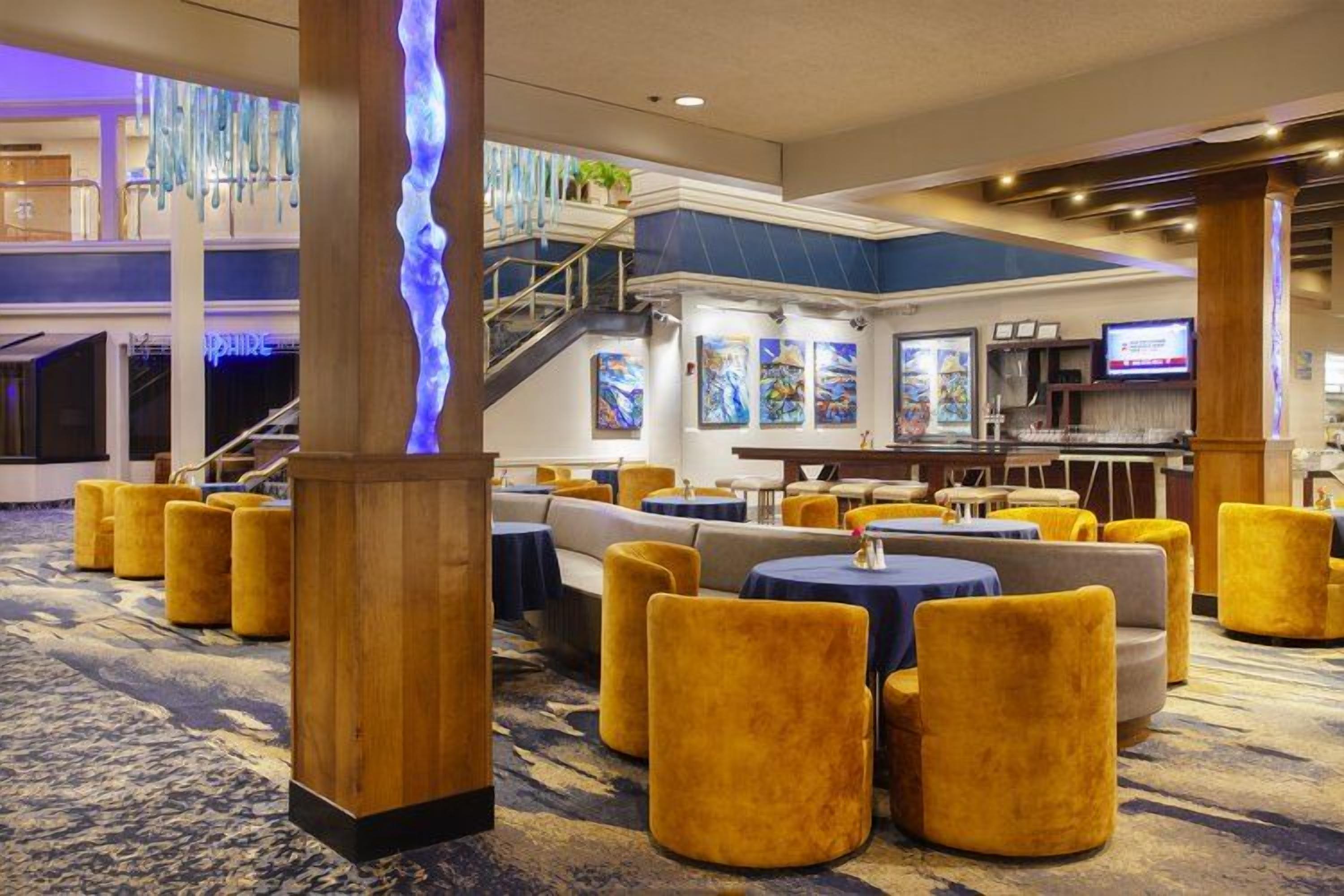 A hotel lobby with a central staircase, seating areas with round tables, and a bar.