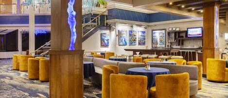 A hotel lobby with a central staircase, seating areas with round tables, and a bar.