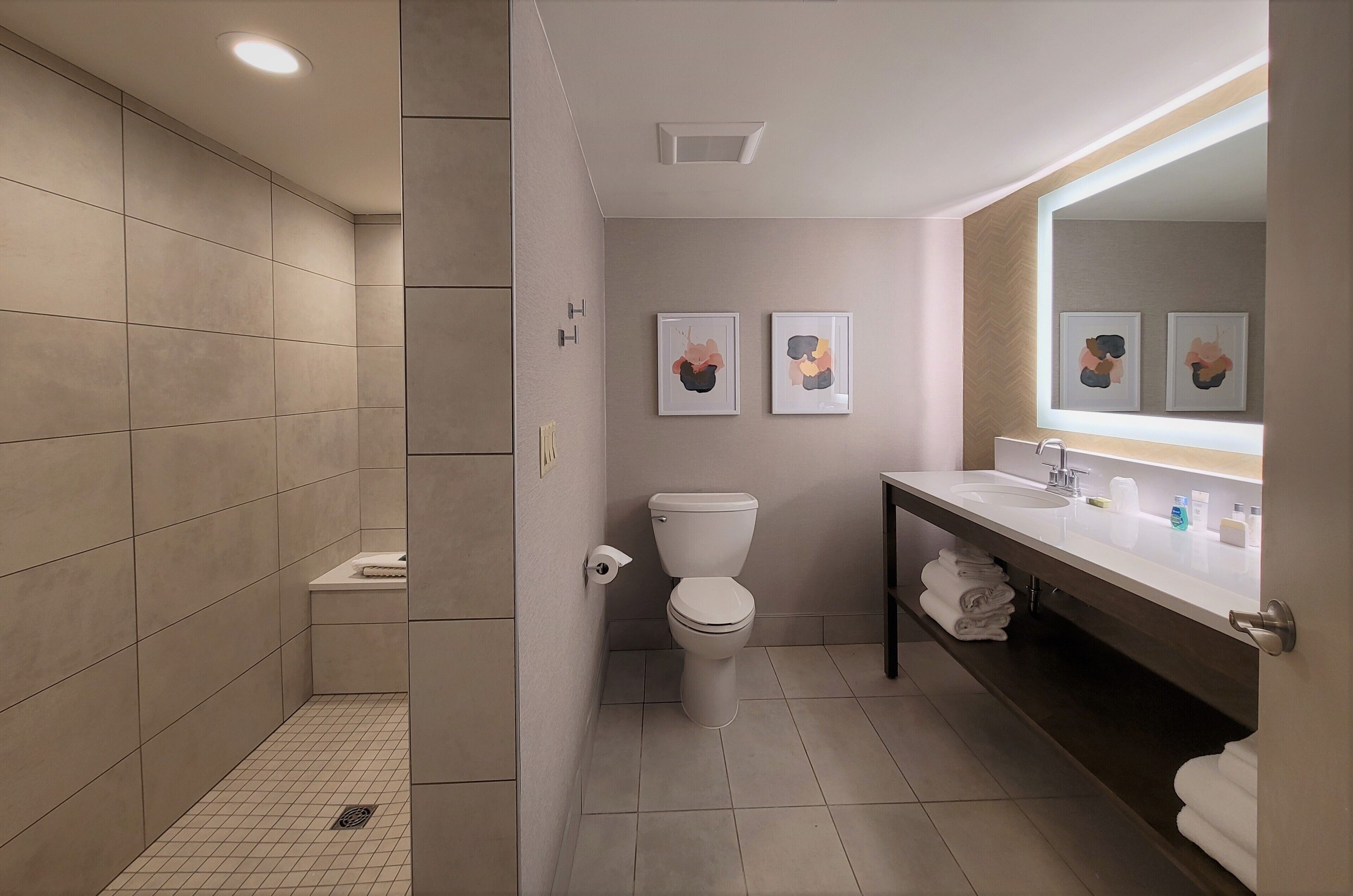 A modern bathroom with a toilet, sink, and shower. There are two framed pictures on the wall, and towels are neatly stacked on the counter.