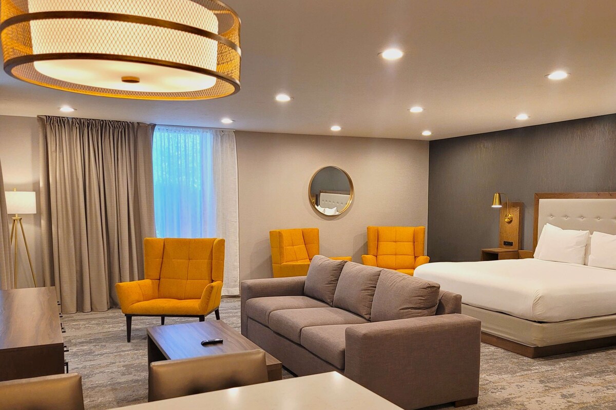 Une chambre d’hôtel comprenant un lit, deux fauteuils, un canapé, un miroir rond, une lampe sur pied et une table de chevet.