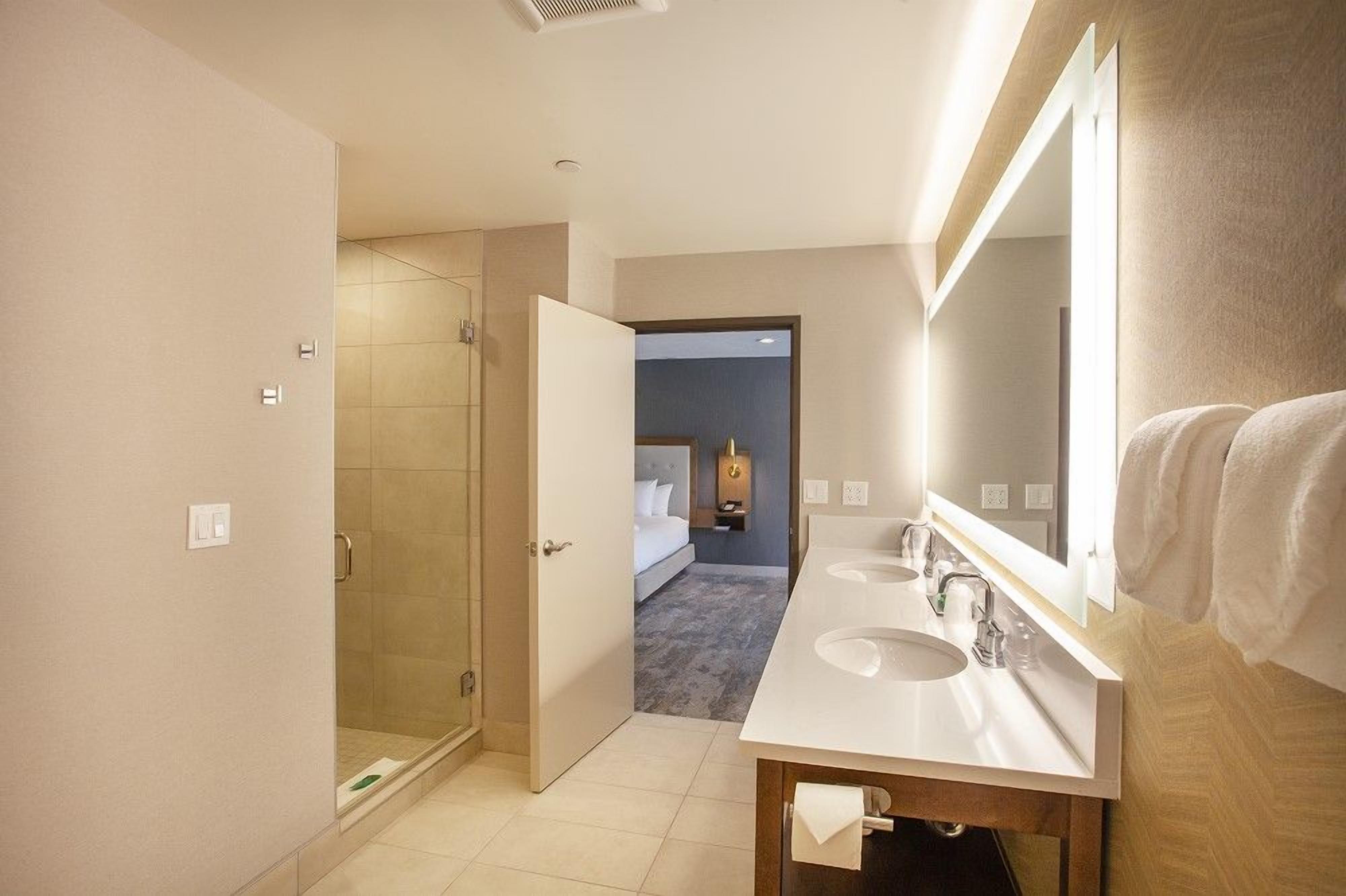 Un baño de hotel con ducha, dos lavamanos, un espejo grande y una cama visible a través de una puerta abierta.