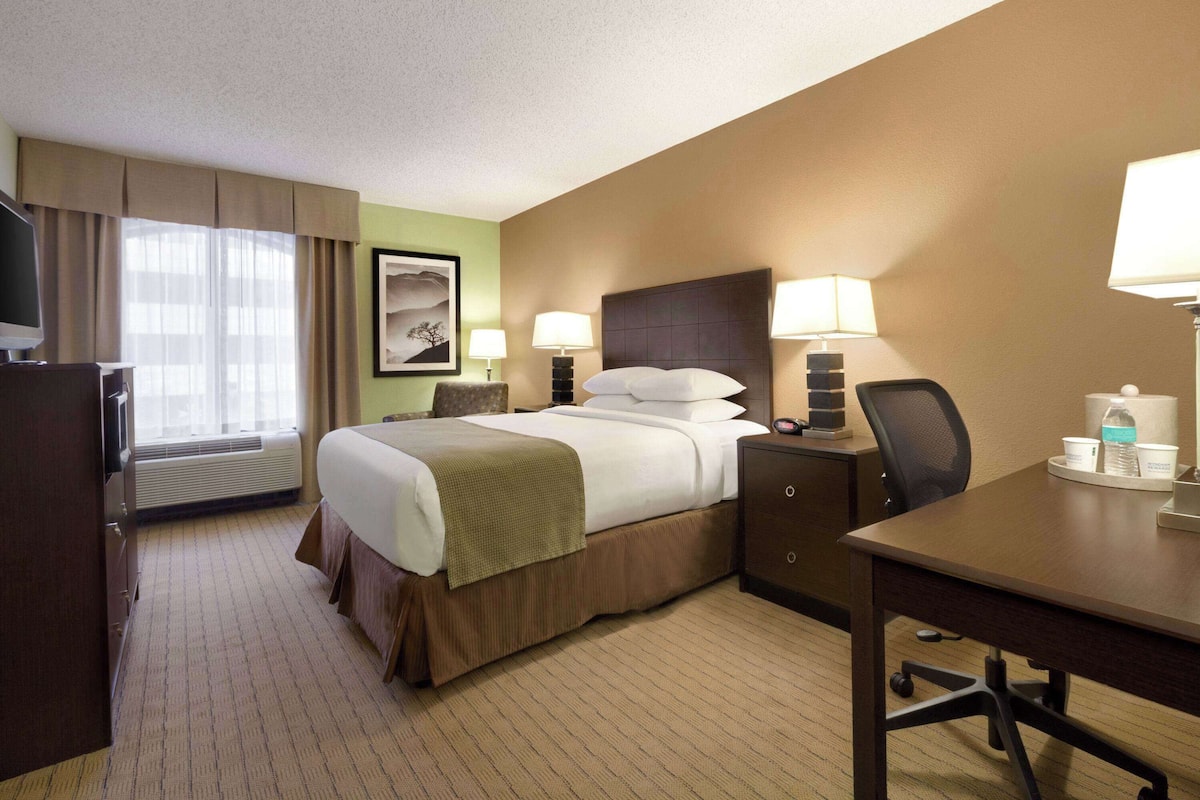 A hotel room with a bed, two bedside tables with lamps, a desk, a chair, a television, and a window with curtains.