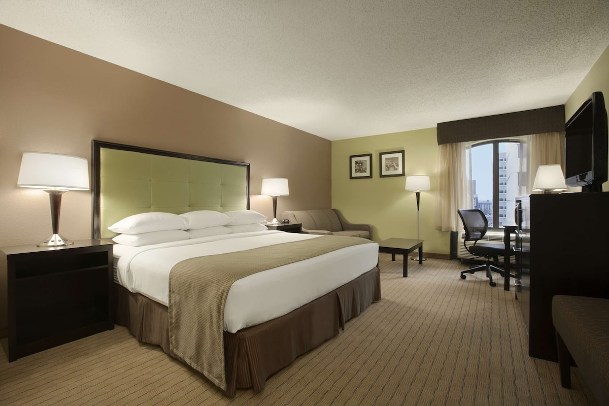 A hotel room with a large bed, two bedside tables with lamps, a desk with a chair, and a television.