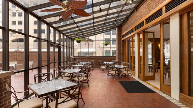 Ein überdachter Patio-Bereich mit Tischen und Stühlen, großen Fenstern und hängenden Pflanzen.