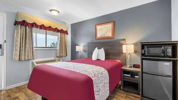 A hotel room with a bed, a microwave, a refrigerator, a nightstand, a lamp, a window with curtains, and a painting on the wall.