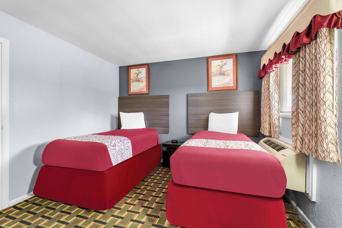 A hotel room with two single beds, a small table, a heater, and a window with curtains.