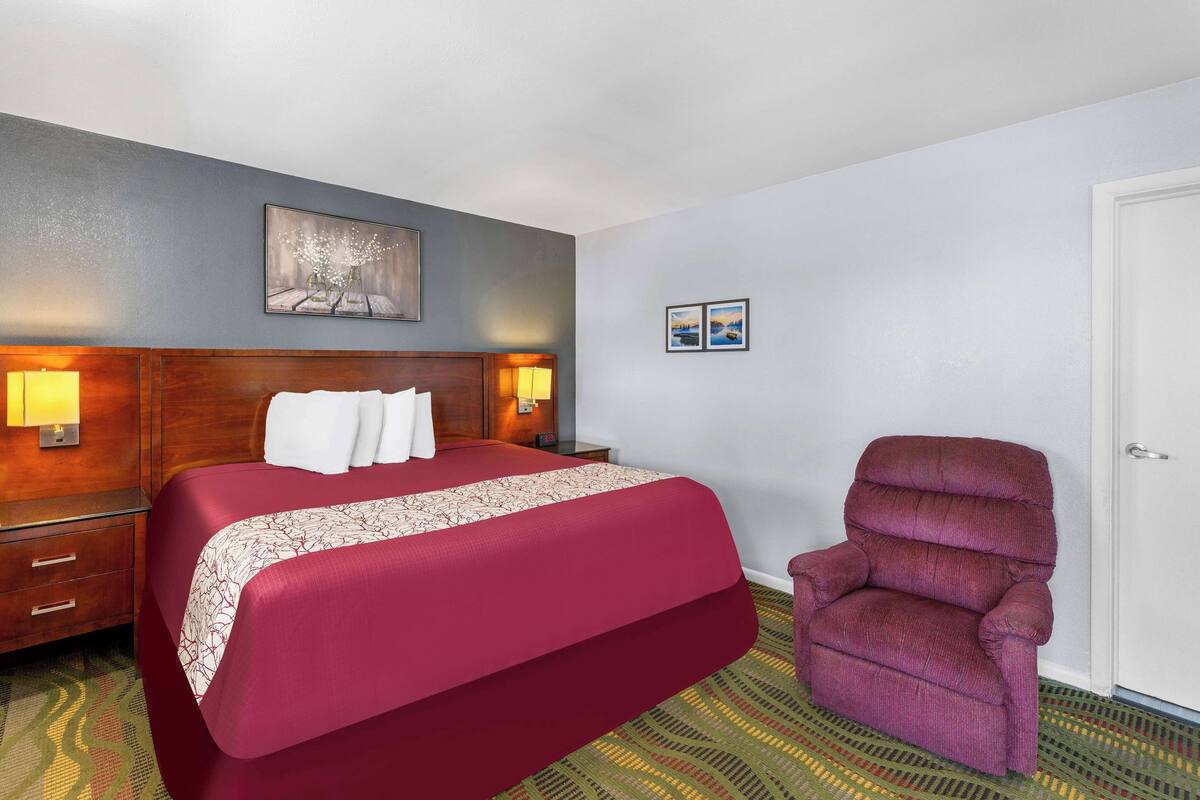 A hotel room with a large bed, wooden headboard, two bedside lamps, a chair, and a door.