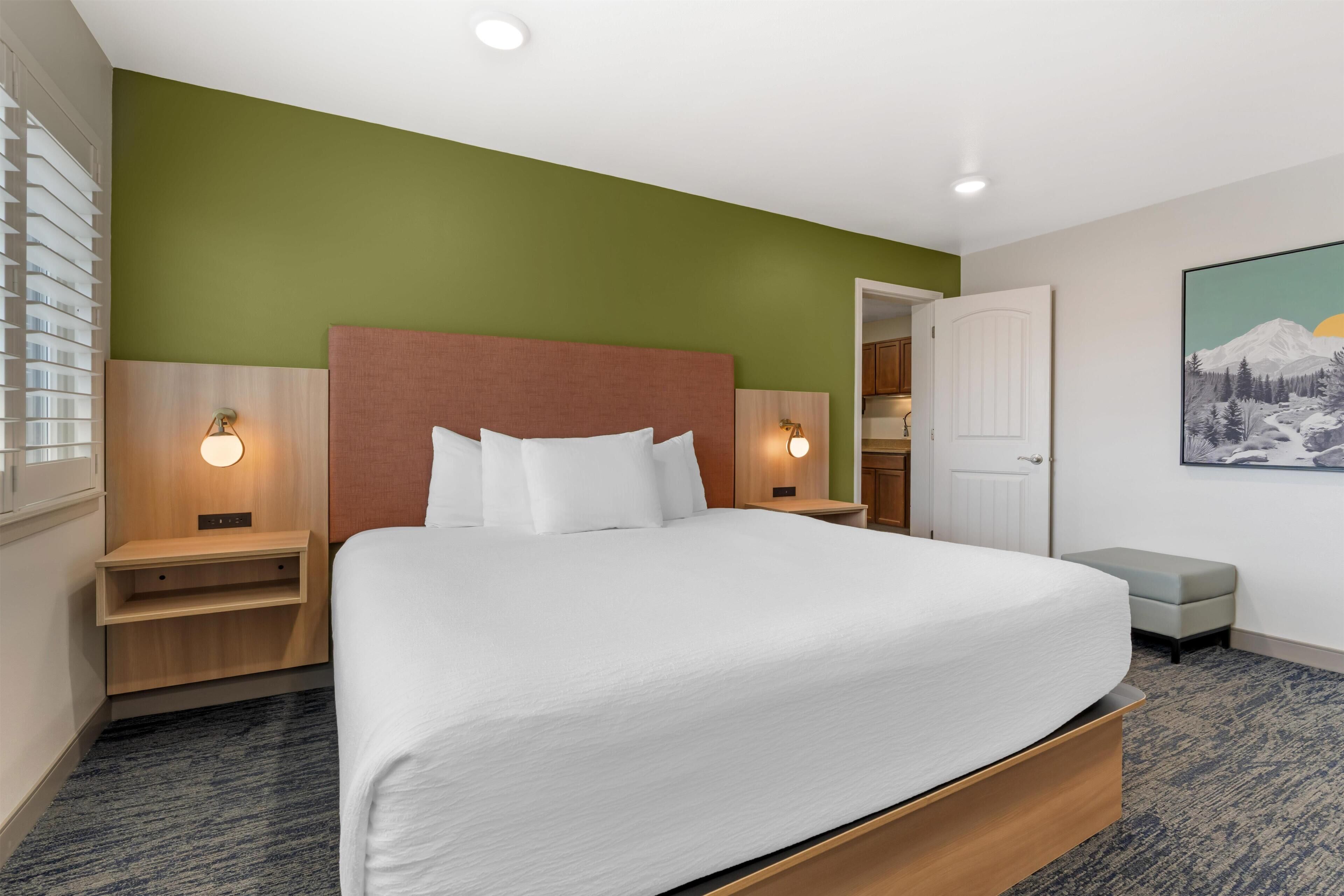 A hotel room with a large bed, bedside tables, a nightstand, a bench, a door, and a painting on the wall.
