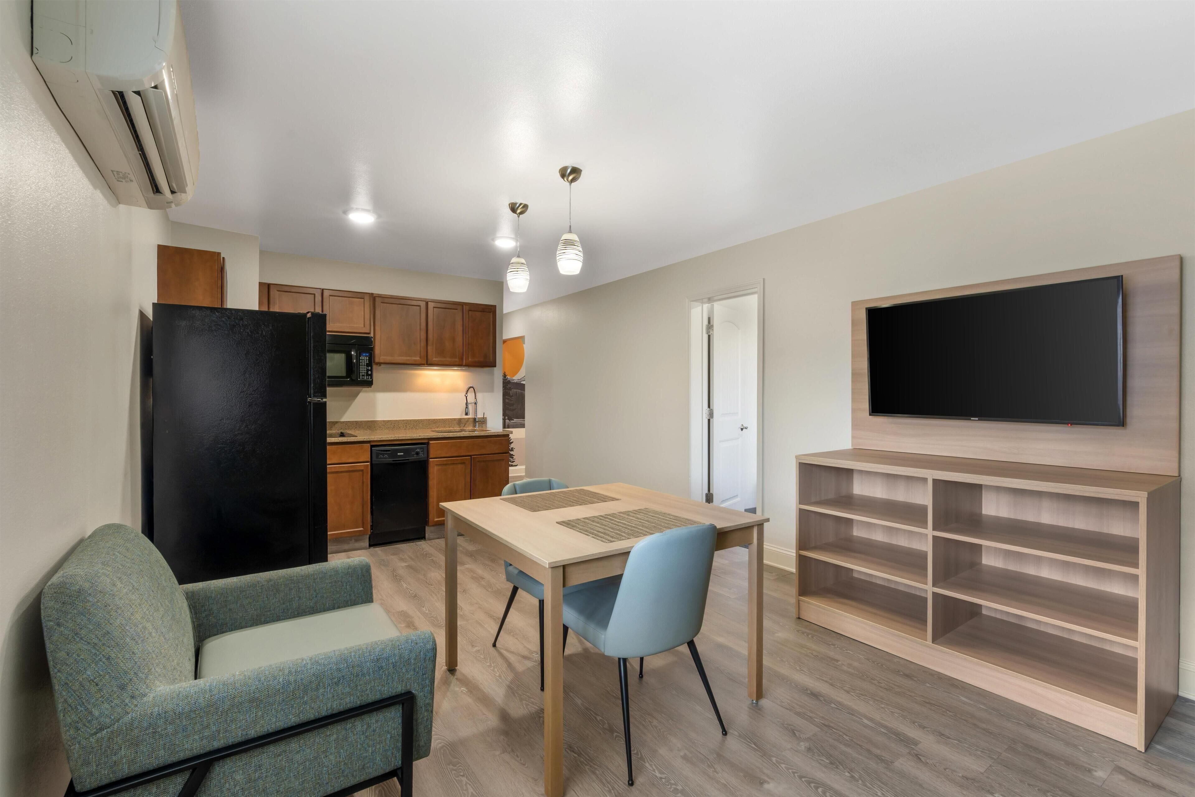 A compact kitchen with a refrigerator, microwave, and wooden cabinets. There is a dining area with a table and chairs, and a TV mounted on the wall.