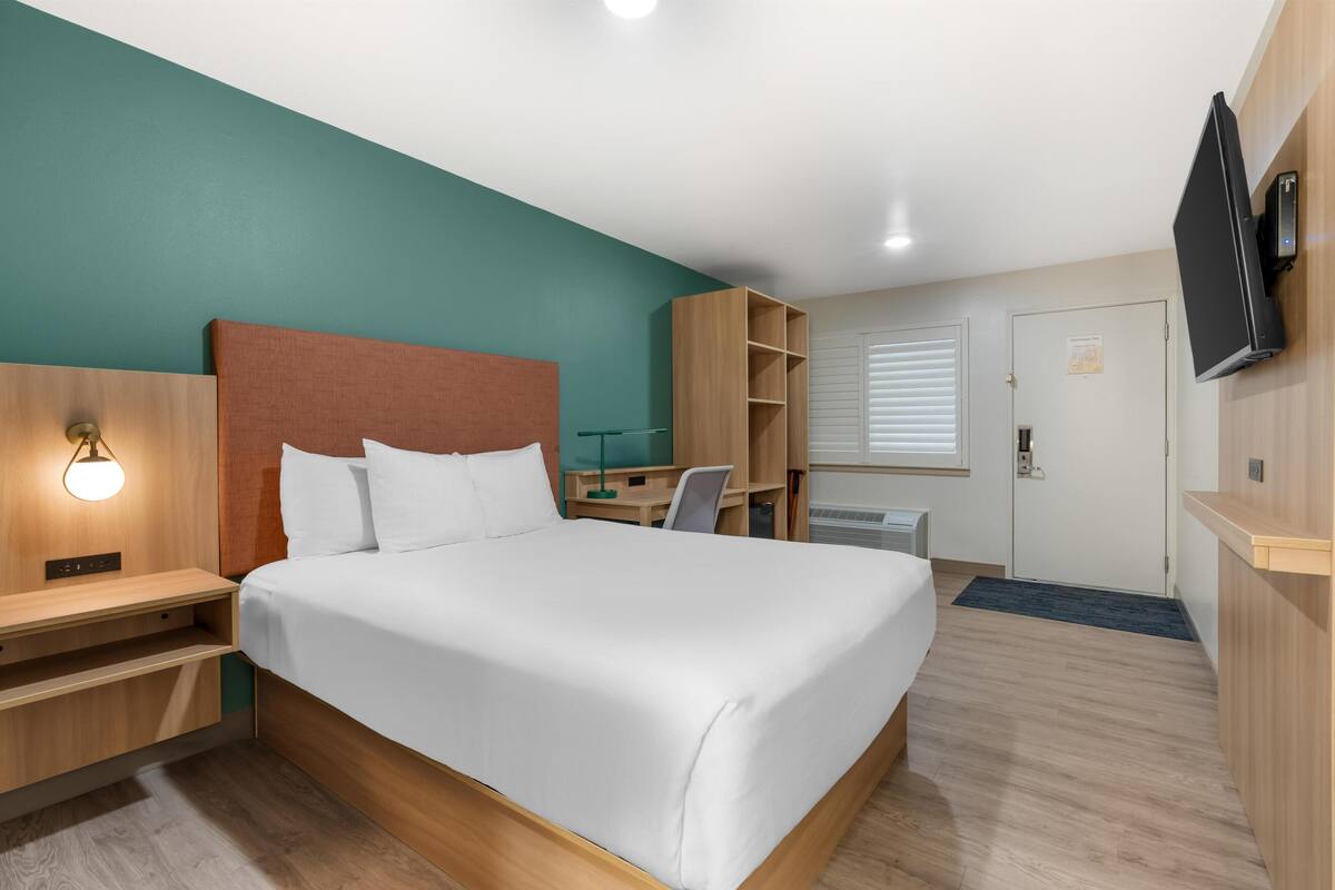 Ett hotellrum med en säng, ett skrivbord, en stol, en TV, en garderob och en dörr.
